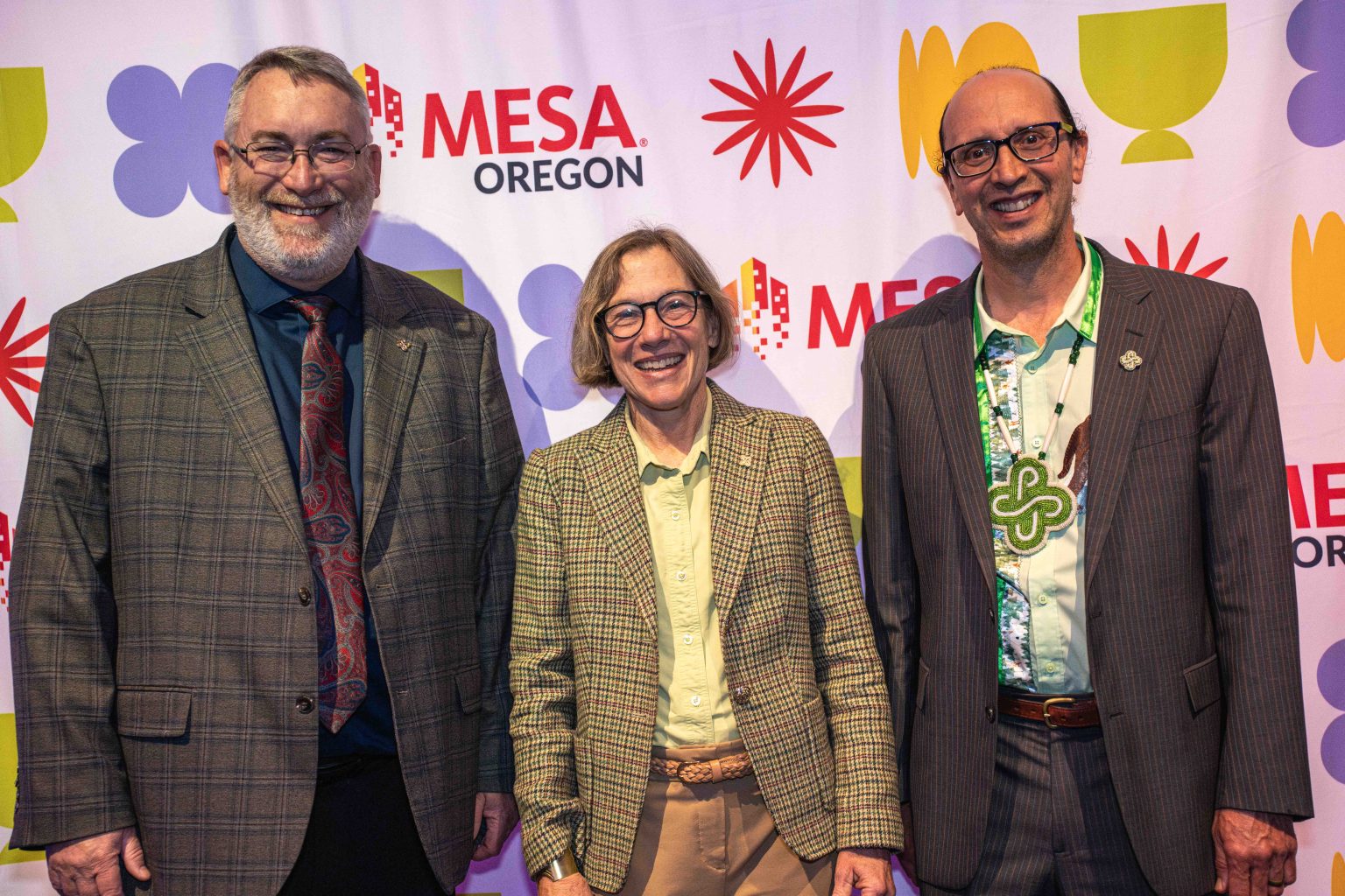 Three adults in business attire smile and pose together in front of a colorful step-and-repeat banner featuring the MESA Oregon logo and abstract shapes at the MESA Through the Decades 2025 Photo Gallery.