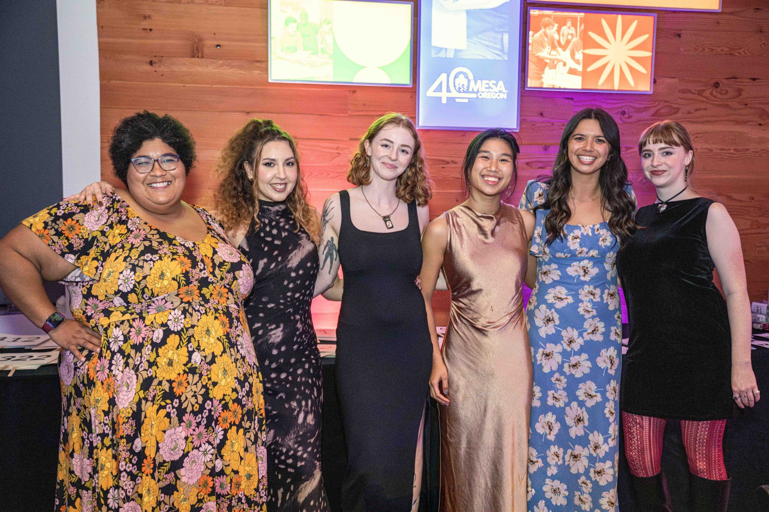 Six women stand together smiling in a row, dressed in colorful dresses, in front of a wooden wall decorated with vibrant posters and a sign reading 40 MESA, capturing the spirit of the MESA Through the Decades 2025 Photo Gallery.