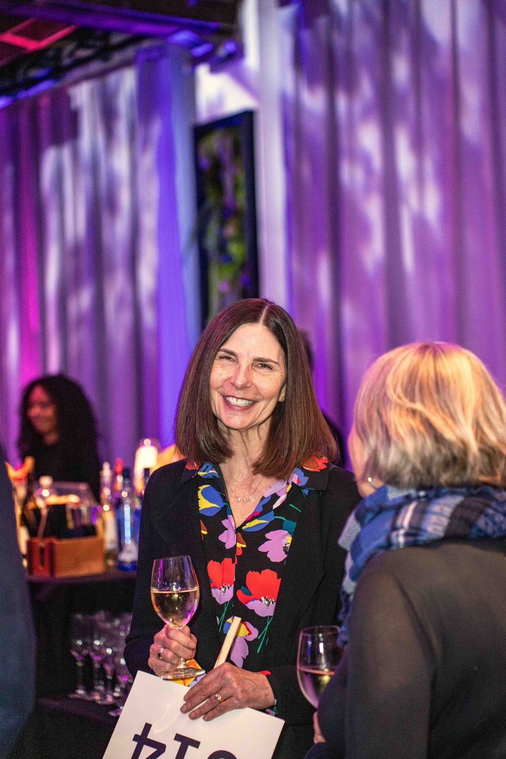 A woman with straight brown hair holding a glass of white wine smiles and talks to another guest at the MESA Through the Decades 2025 Photo Gallery, set at an indoor event with purple lighting and a bar in the background.