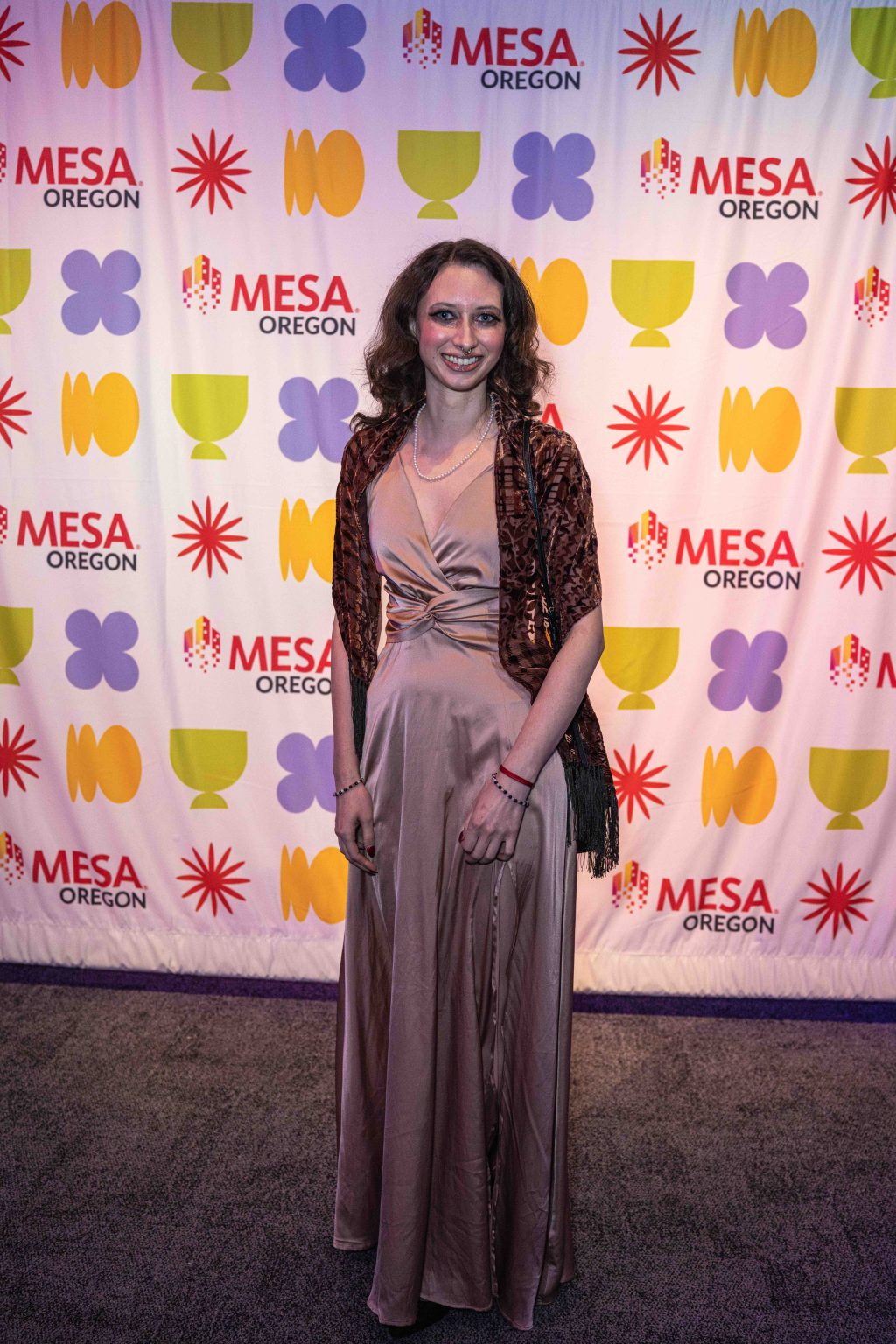 A woman in a long, silky beige dress and patterned shawl smiles while standing before a colorful step and repeat banner with MESA Oregon logos, geometric shapes, and the MESA Through the Decades 2025 Photo Gallery backdrop.