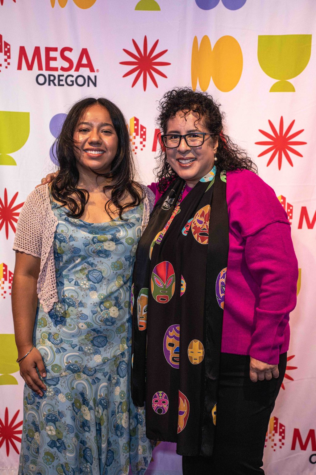 Two women stand smiling in front of a colorful backdrop with the text MESA Oregon and various shapes, captured for the MESA Through the Decades 2025 Photo Gallery. One wears a blue floral dress; the other, glasses, a magenta sweater, and a mask-patterned scarf.