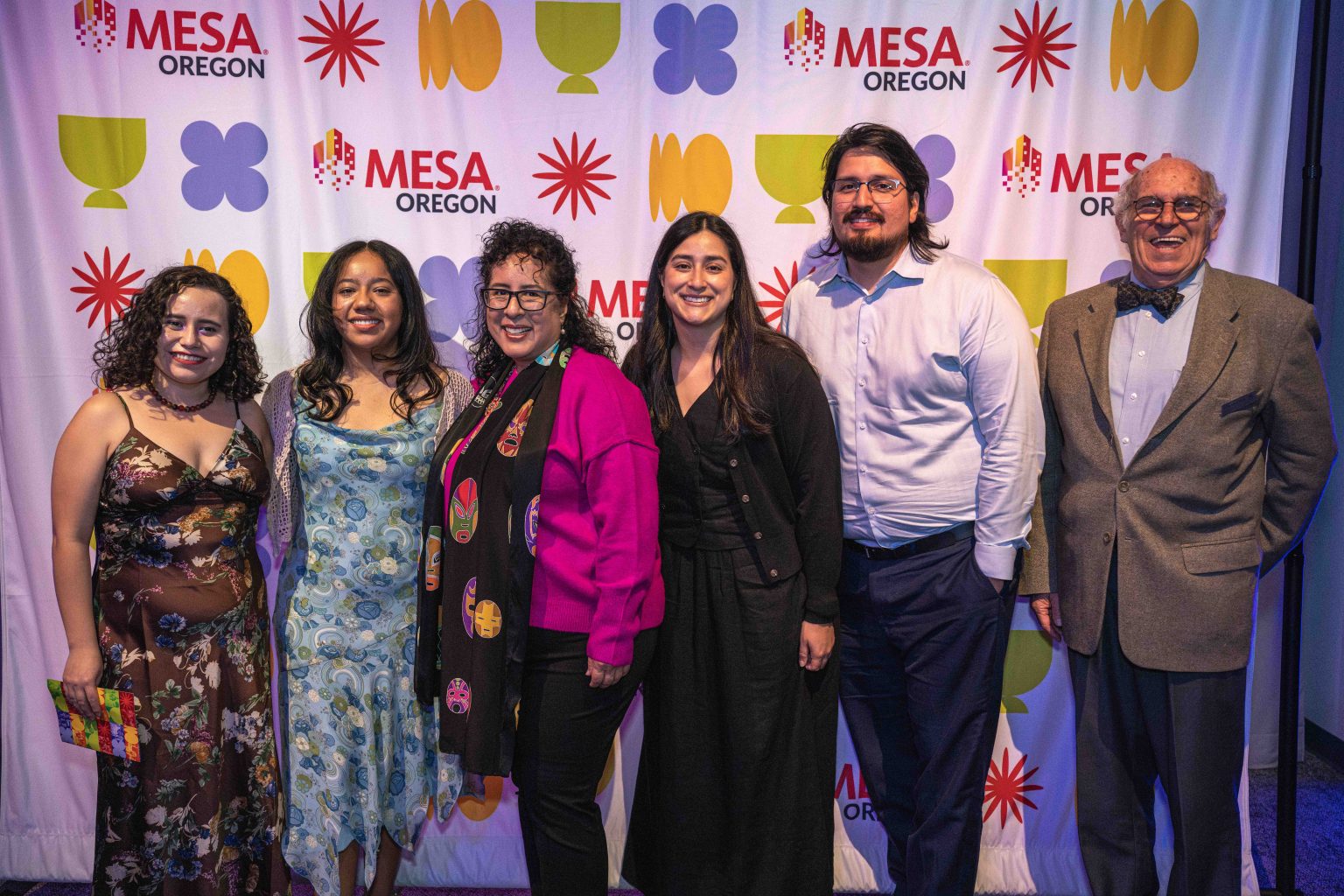 Six people stand smiling in front of a step-and-repeat banner with the MESA Oregon logo and colorful symbols, dressed in formal and semi-formal attire for the MESA Through the Decades 2025 Photo Gallery.