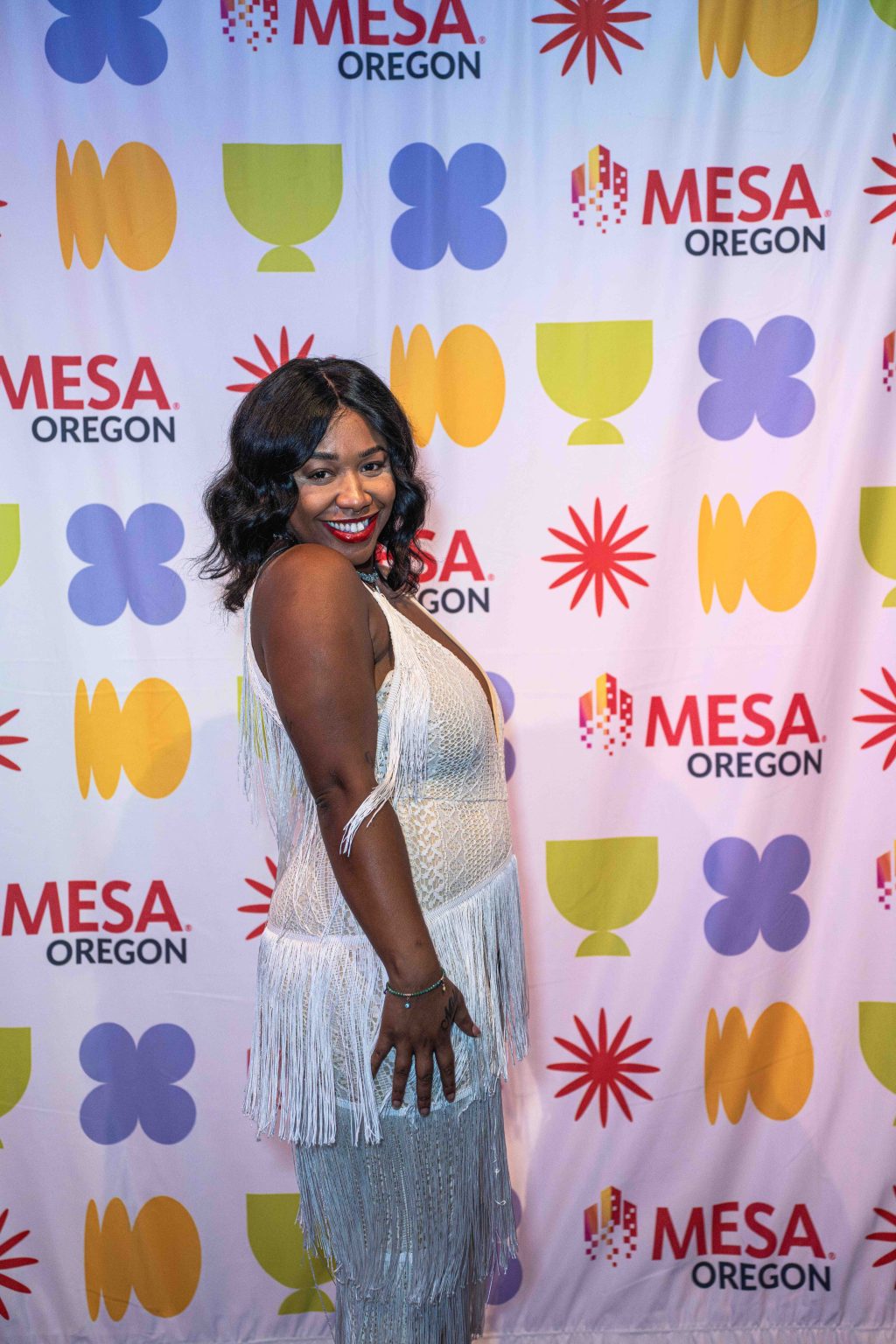 A woman in a white fringed dress poses and smiles in front of a step-and-repeat banner with colorful shapes and MESA OREGON logos at the MESA Through the Decades 2025 Photo Gallery.
