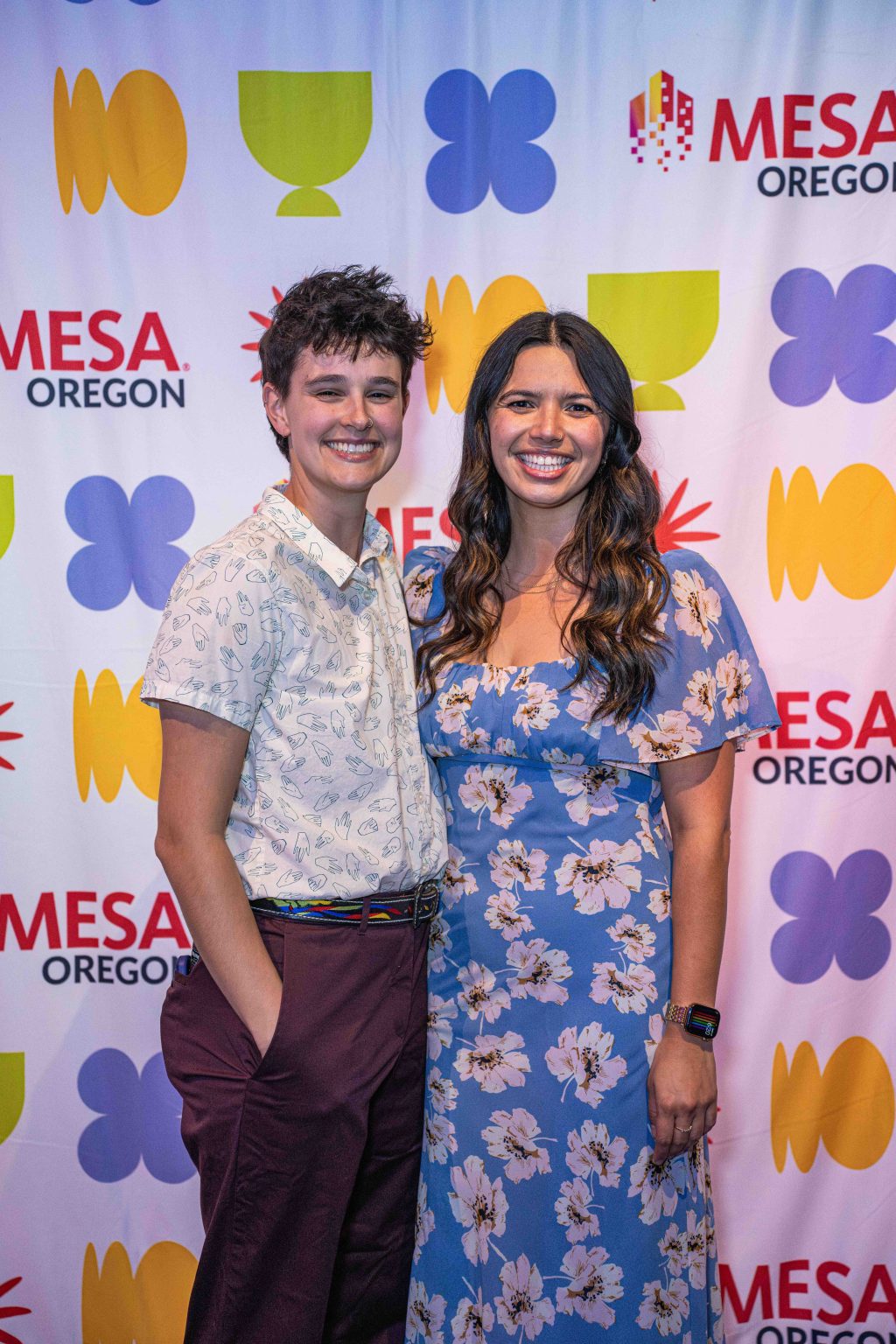 Two people smile in front of a step-and-repeat backdrop with colorful shapes and MESA Oregon logos at the MESA Through the Decades 2025 Photo Gallery. One wears a floral dress, the other a patterned shirt and maroon pants.