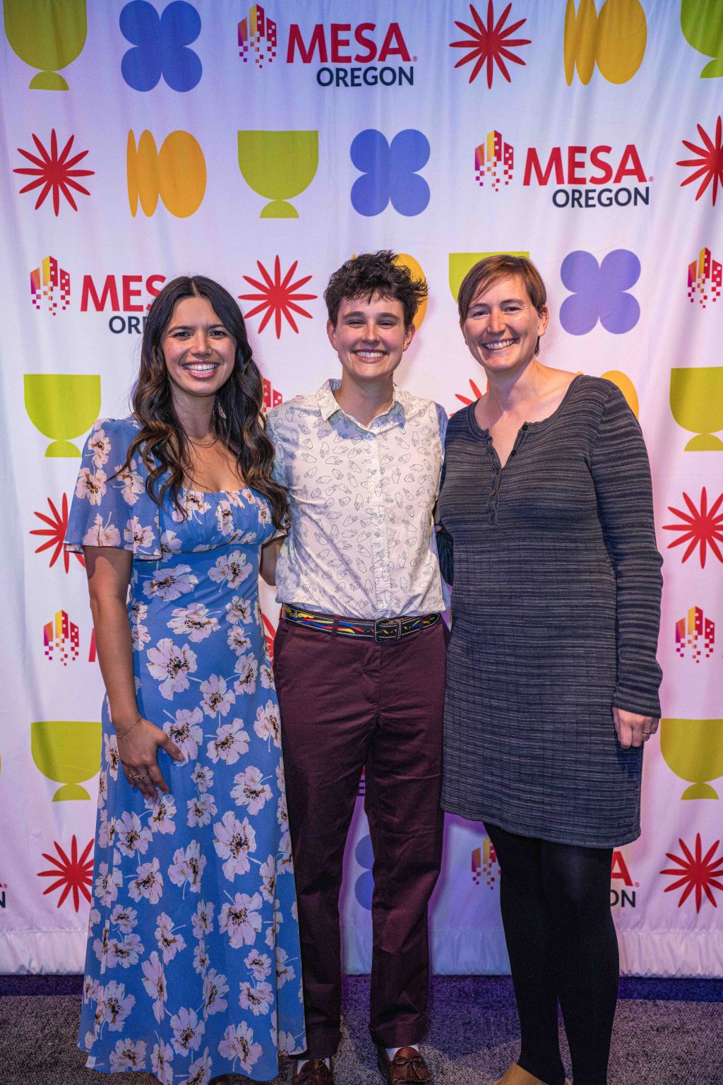 Three people stand smiling in front of a colorful step-and-repeat banner with the MESA Oregon logo and various shapes at the MESA Through the Decades 2025 Photo Gallery. Each is dressed in vibrant attire, capturing the spirit of the event.
