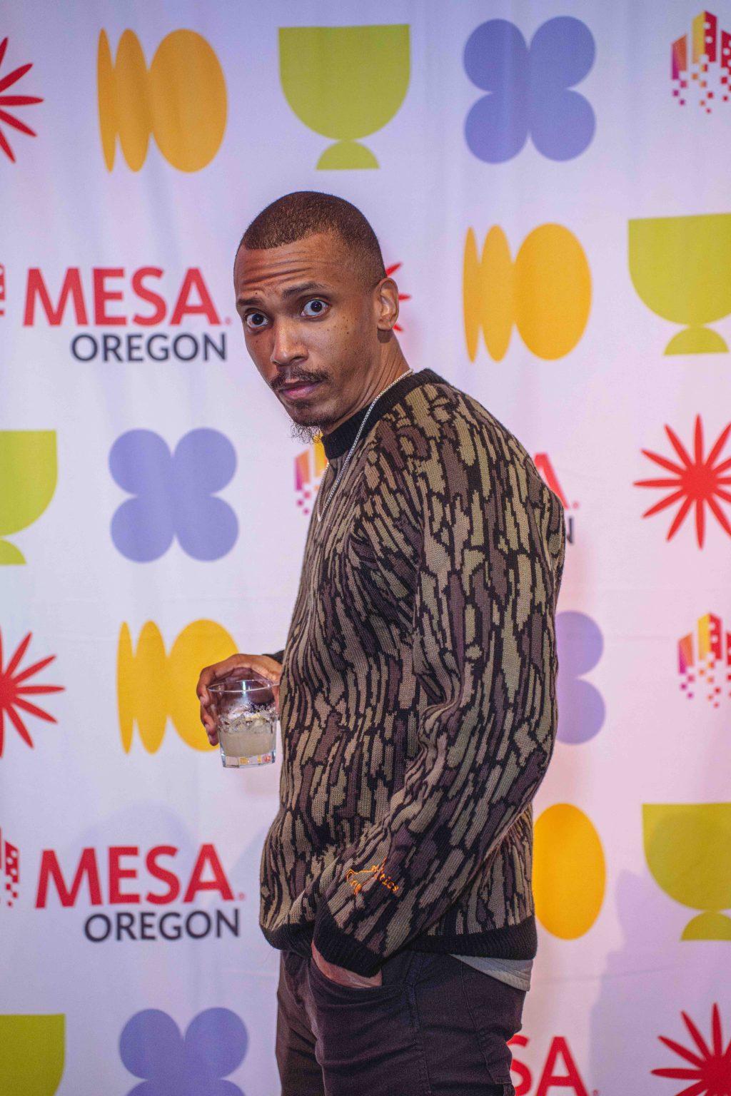 A man in a patterned sweater holds a drink and looks at the camera while standing in front of a colorful backdrop with abstract shapes and the words MESA OREGON, featured in the MESA Through the Decades 2025 Photo Gallery.