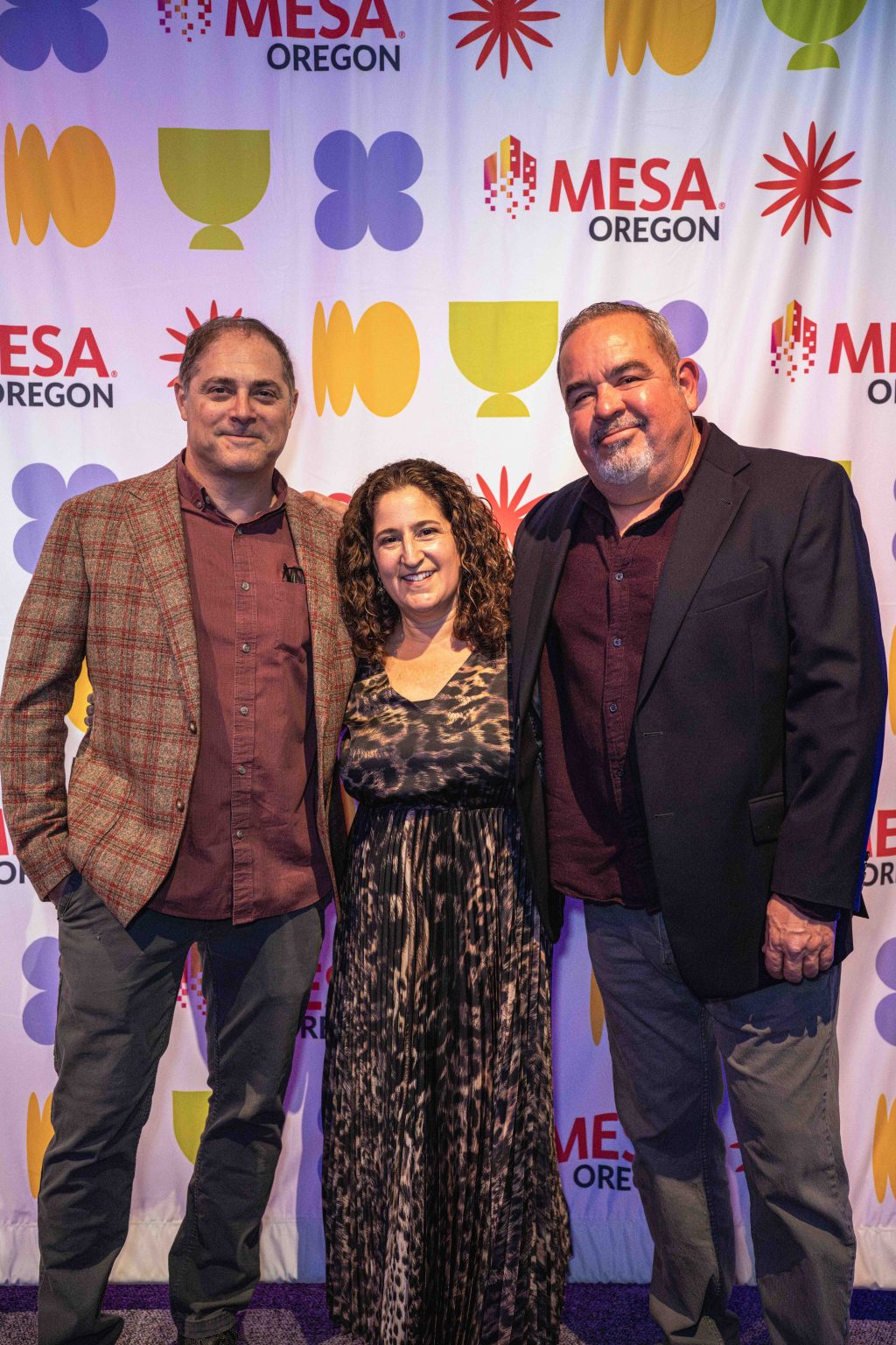 Three adults, two men and one woman, stand smiling together in front of a colorful backdrop with MESA OREGON logos and geometric shapes at the MESA Through the Decades 2025 Photo Gallery event.