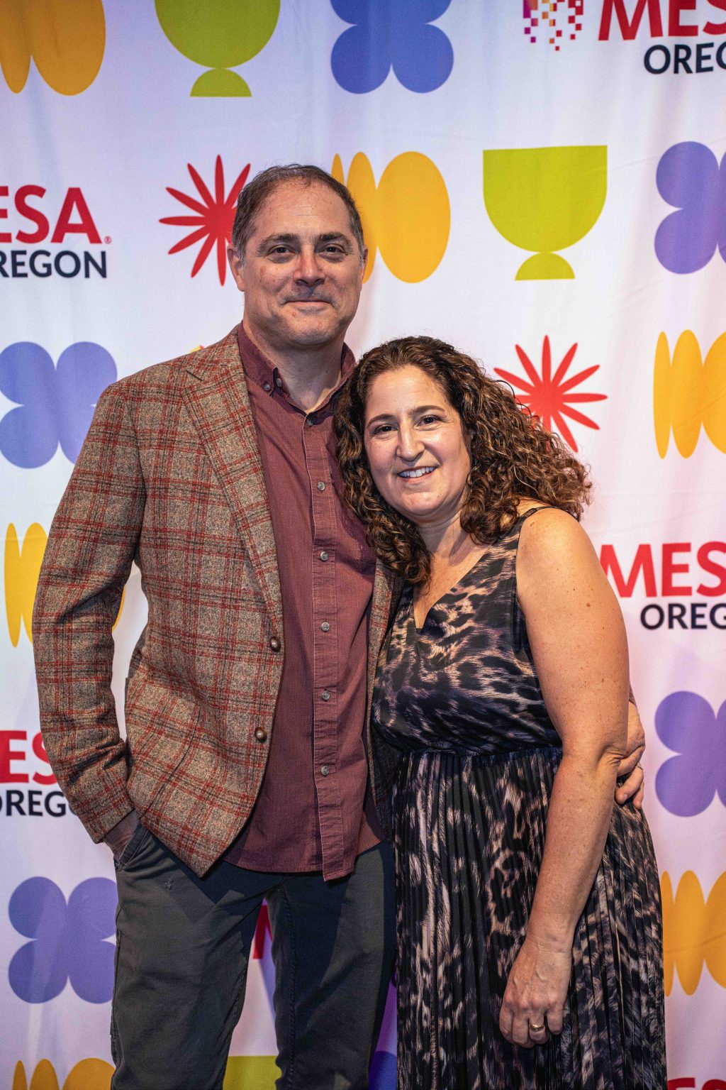 A man in a plaid jacket and maroon shirt stands next to a woman in a patterned sleeveless dress. Smiling, they pose together in front of the colorful MESA Oregon backdrop at the MESA Through the Decades 2025 Photo Gallery.
