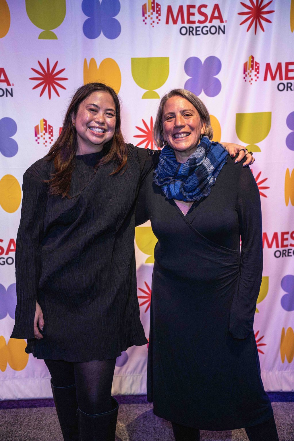 Two women stand smiling with arms around each other in front of a colorful MESA Oregon backdrop featuring geometric shapes and the MESA Oregon logo, capturing a joyful moment for the MESA Through the Decades 2025 Photo Gallery.