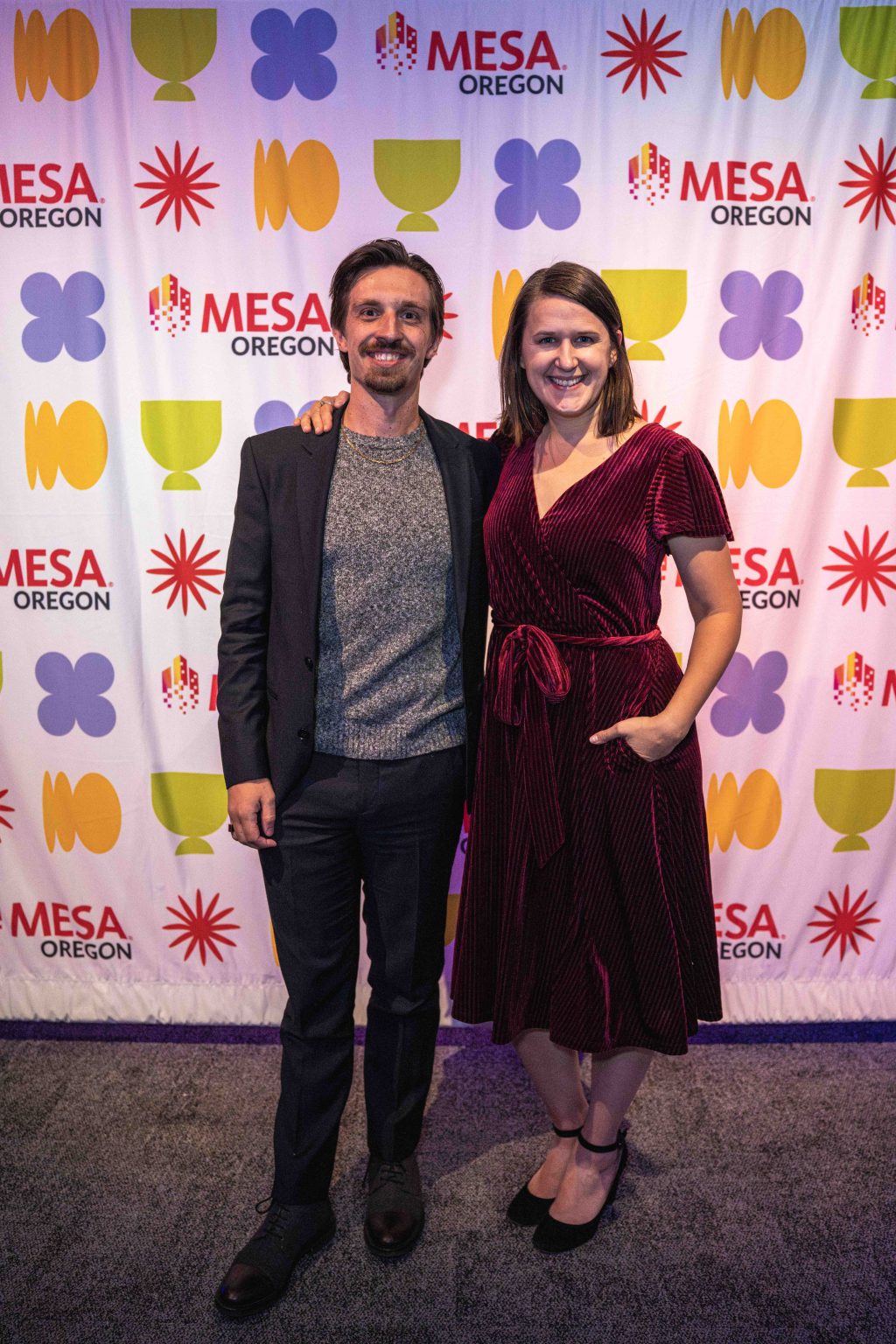 A man in a suit and a woman in a red dress stand smiling together in front of a colorful step-and-repeat backdrop with MESA OREGON logos and abstract shapes at the MESA Through the Decades 2025 Photo Gallery.