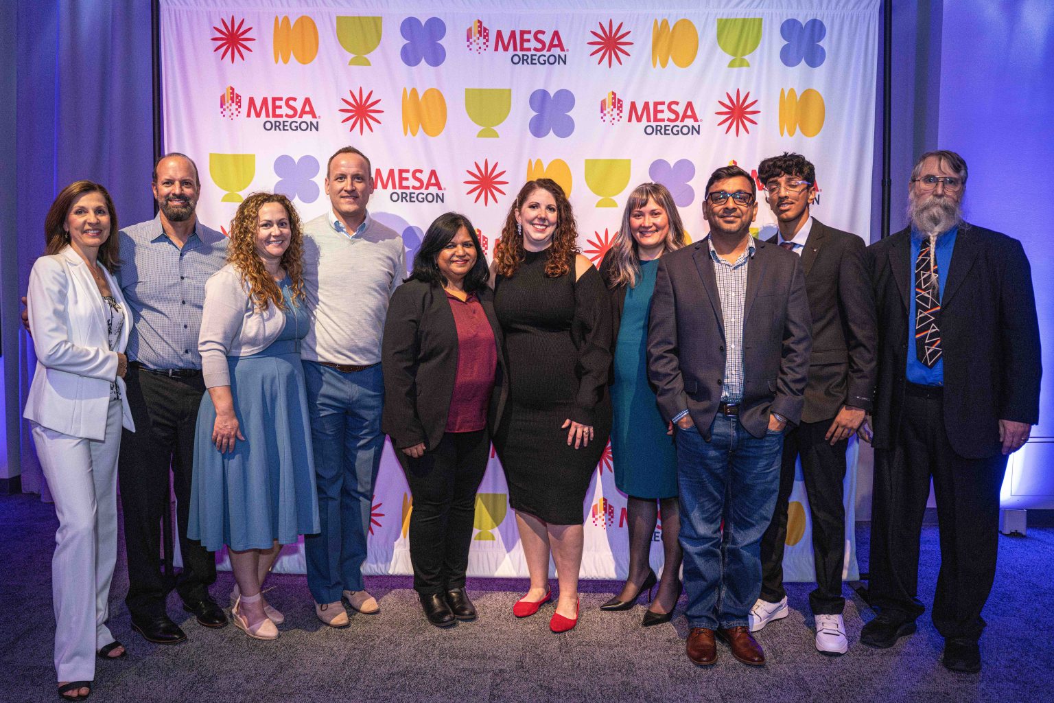 A group of eleven people pose together, smiling in front of a colorful Mesa Oregon backdrop for the MESA Through the Decades 2025 Photo Gallery. The group is dressed in business and semi-formal attire.
