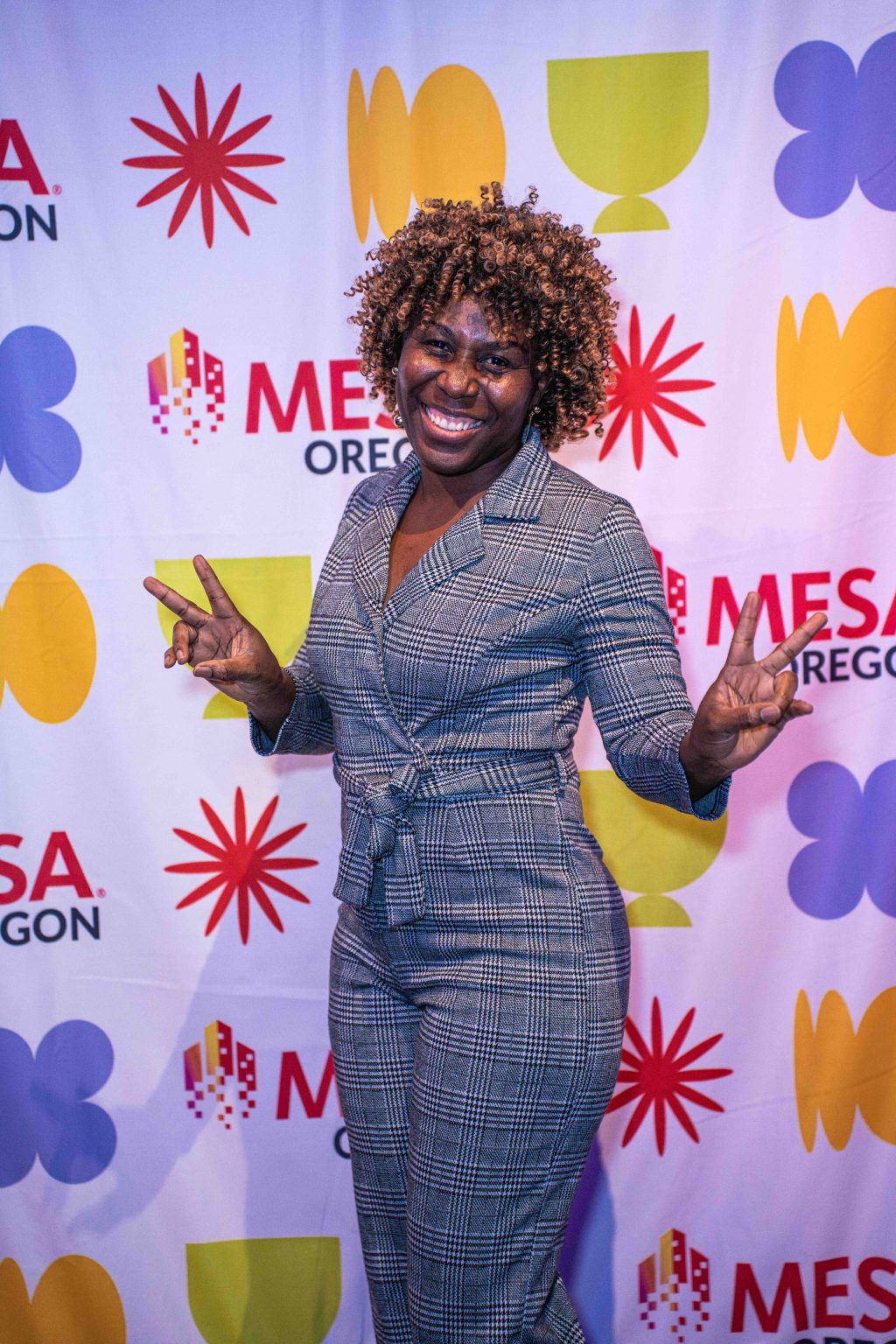 A smiling woman in a grey plaid jumpsuit flashes double peace signs while standing in front of the colorful MESA Through the Decades 2025 Photo Gallery step-and-repeat banner.