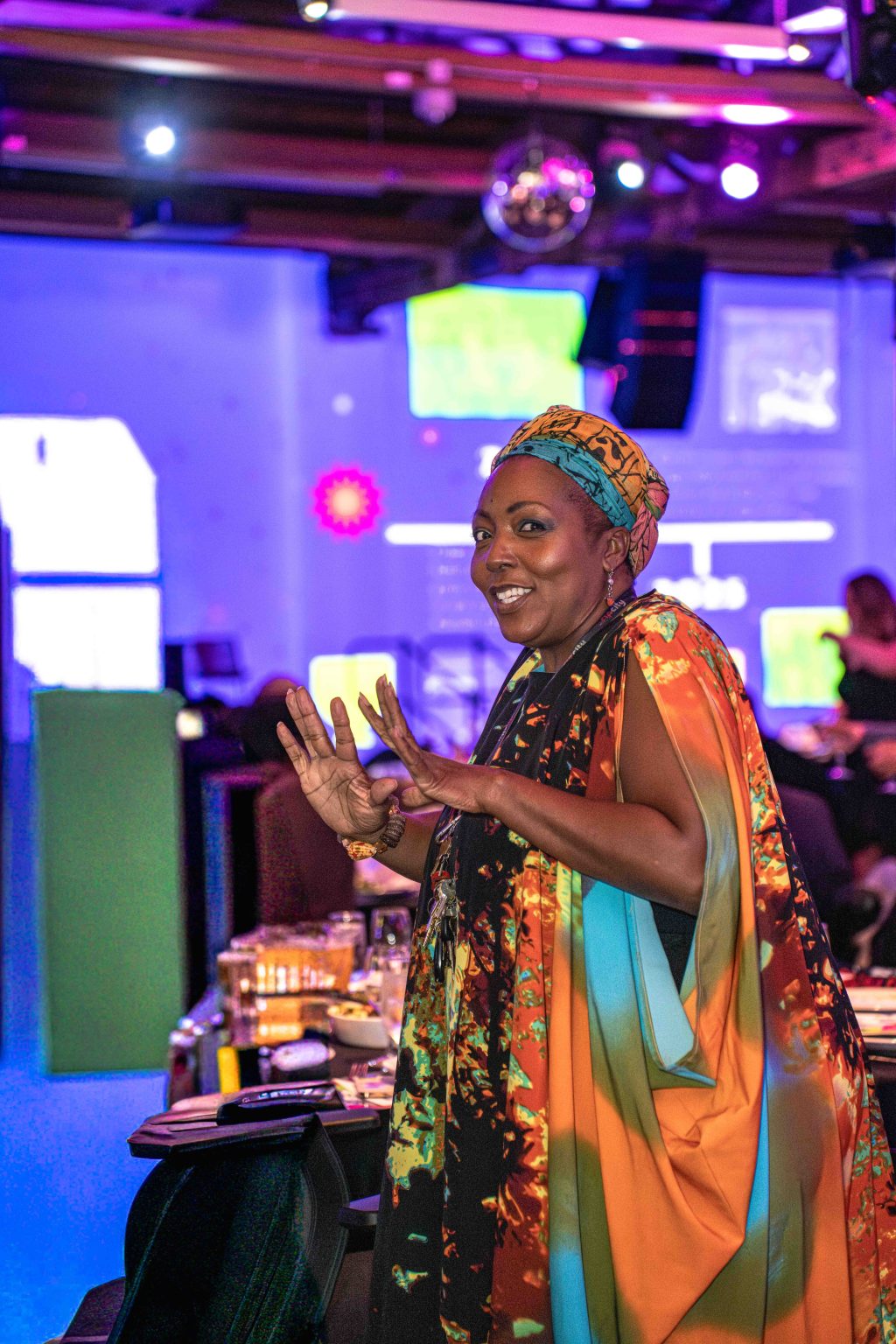 A woman in a colorful dress and head wrap smiles and gestures with her hands at the MESA Through the Decades 2025 Photo Gallery, an indoor event with purple lighting and people seated at tables in the background.