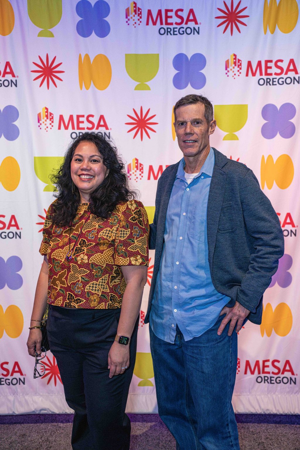 Two people stand smiling in front of a colorful step-and-repeat banner with the words MESA Oregon and abstract shapes. This vibrant moment from the MESA Through the Decades 2025 Photo Gallery shows one in a patterned shirt, the other in a blue jacket.
