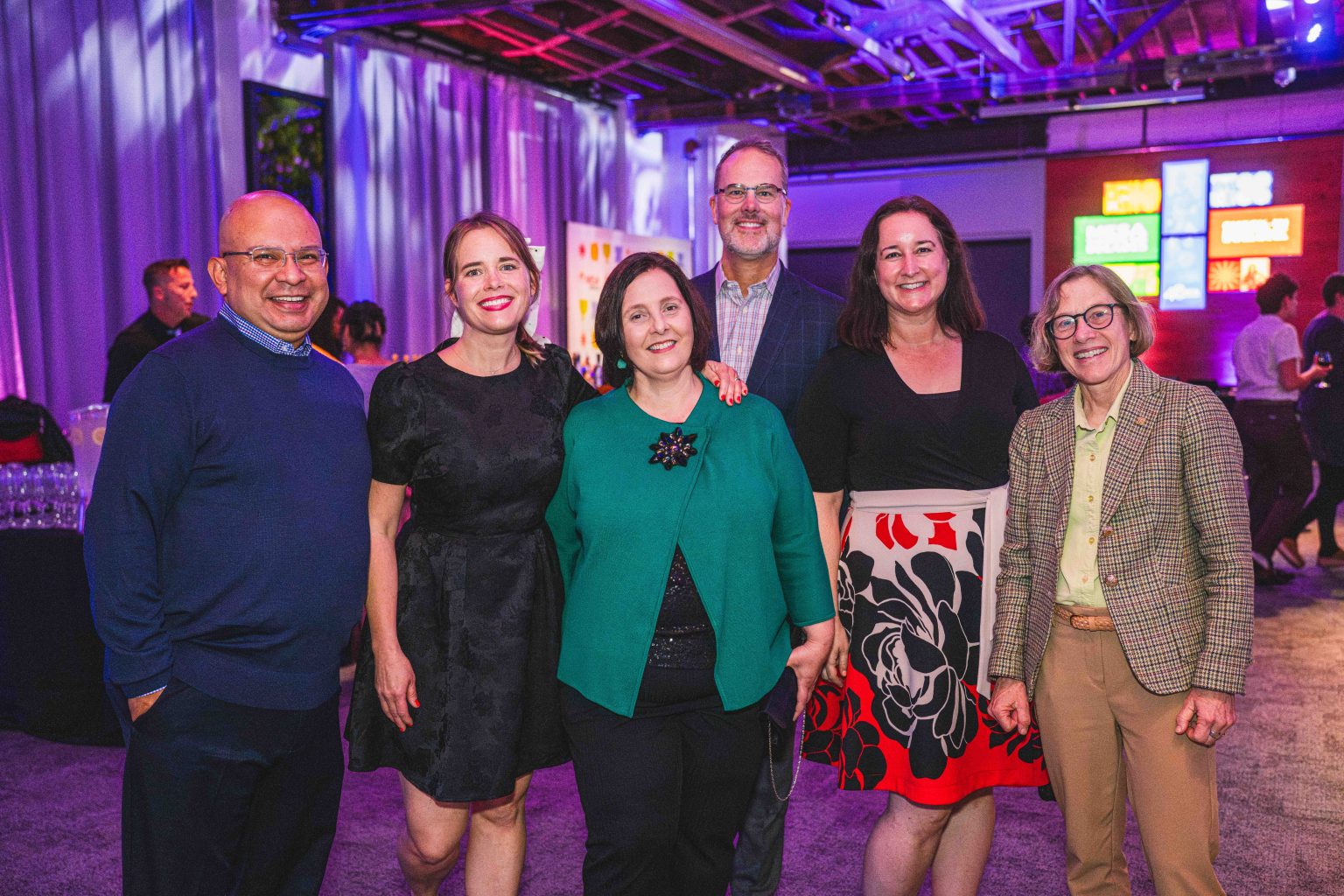 Six adults, three men and three women, stand together smiling for a group photo at the MESA Through the Decades 2025 Photo Gallery event, with colorful lighting and neon signs glowing in the background.