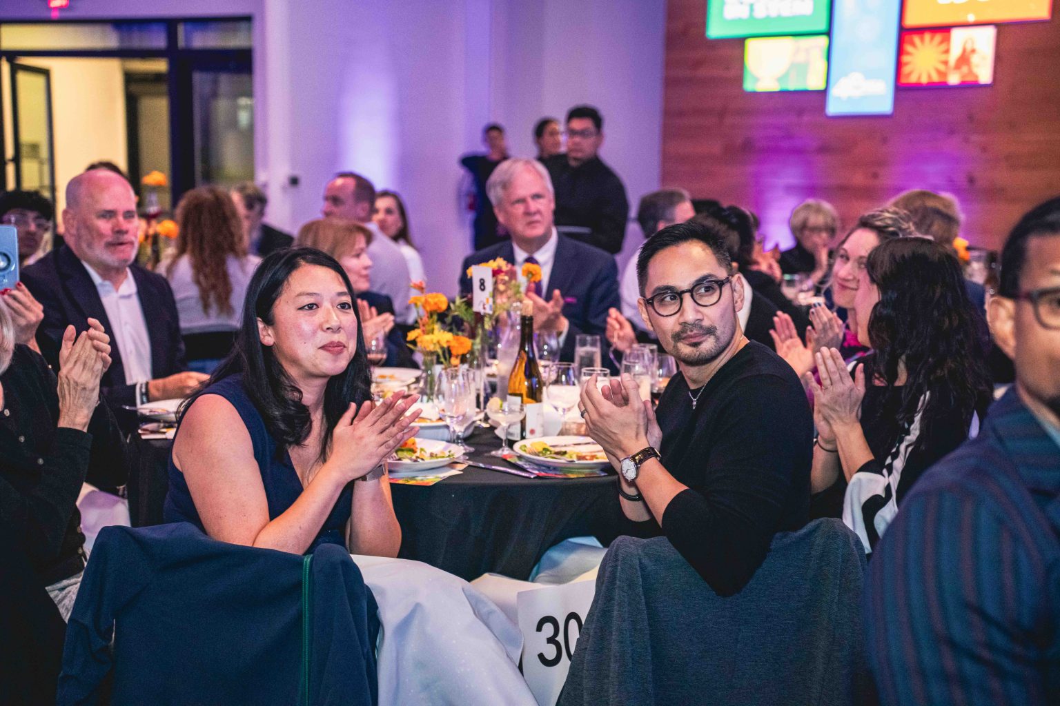 People in formal attire sit at round tables, applauding during the lively MESA Through the Decades 2025 event. Tables are set with food and flowers, while colorful signs adorn a wooden wall—perfect for the Photo Gallery.