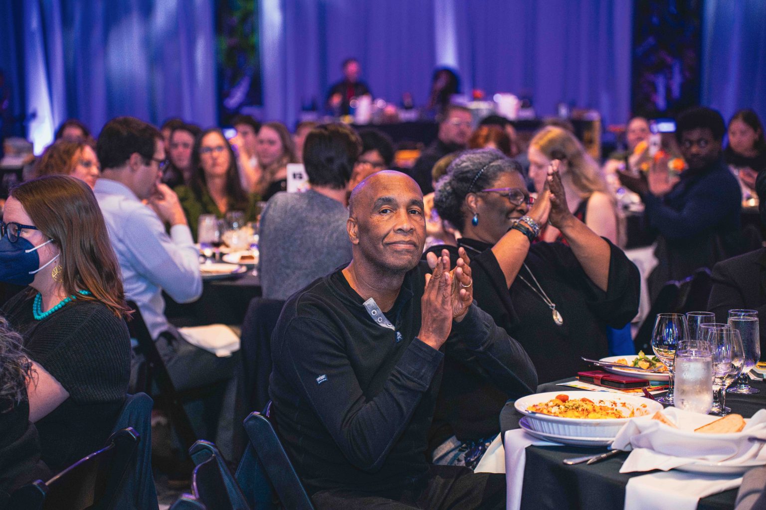 A group of people sit at round tables, dining and applauding during the formal MESA Through the Decades 2025 Photo Gallery event, with subdued blue-purple lighting and plates of food and drinks on the tables.