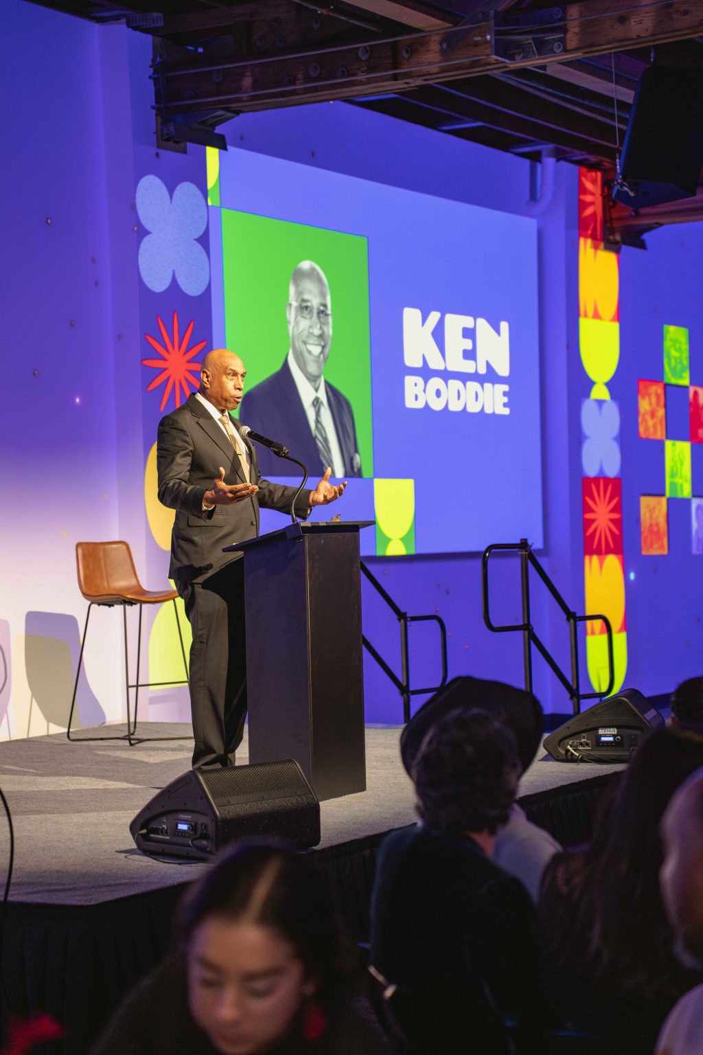 A man in a suit speaks at a podium on stage with a projected image and the text KEN BODDIE behind him. The colorful room, part of the MESA Through the Decades 2025 Photo Gallery, is filled with people seated in the audience.