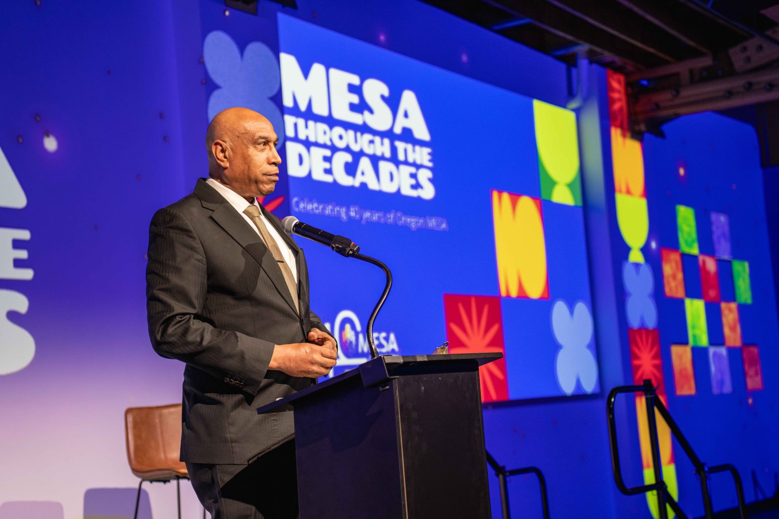 A man in a suit stands at a podium, speaking at an event. Behind him, a large screen reads “MESA THROUGH THE DECADES: Celebrating 45 years of Oregon MESA” with colorful geometric designs from the MESA Through the Decades 2025 Photo Gallery.