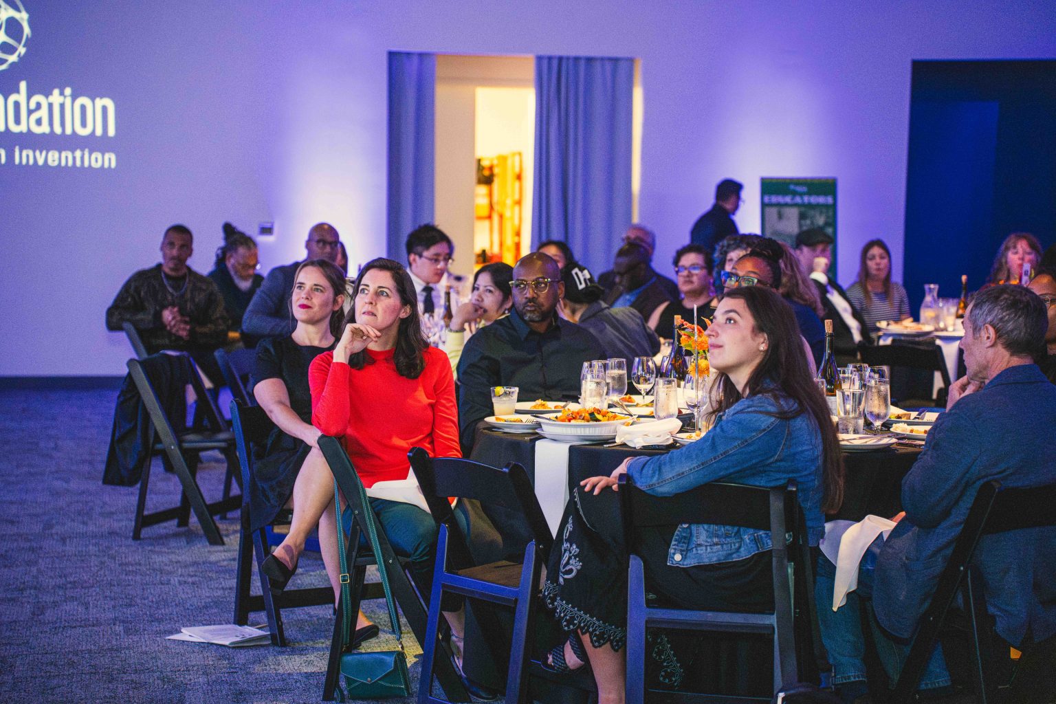 A group of people sit at round tables in a dimly lit event space, attentively facing forward. The tables have plates, glasses, and flower centerpieces. A projected MESA Through the Decades 2025 Photo Gallery logo is visible on the wall.