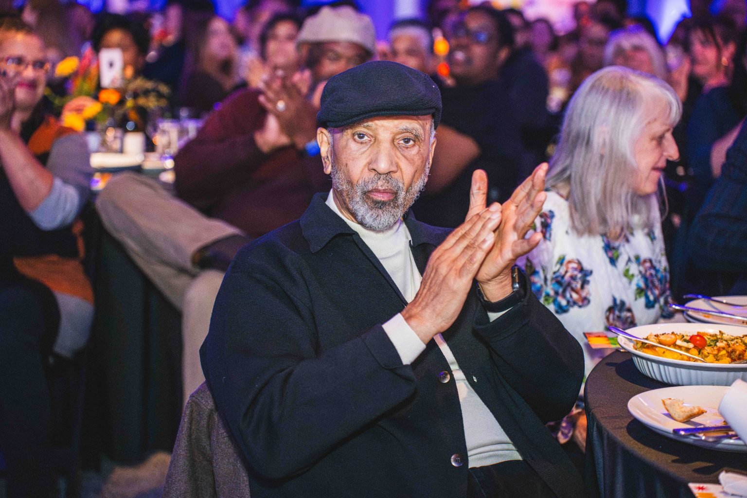 An older man wearing a black cap and jacket sits at a table, clapping at the lively MESA Through the Decades 2025 Photo Gallery event. Plates of food are visible in the foreground, surrounded by a festive crowd.