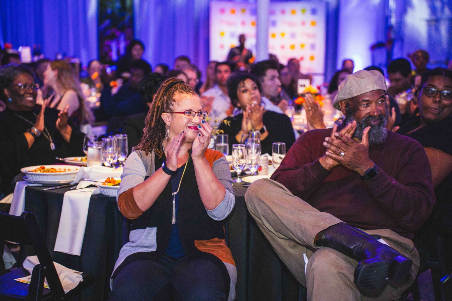 A diverse group of people at the MESA Through the Decades 2025 Photo Gallery sit at round tables with white tablecloths, clapping and smiling during an indoor event with blue lighting. Some are eating, and everyone looks engaged and cheerful.
