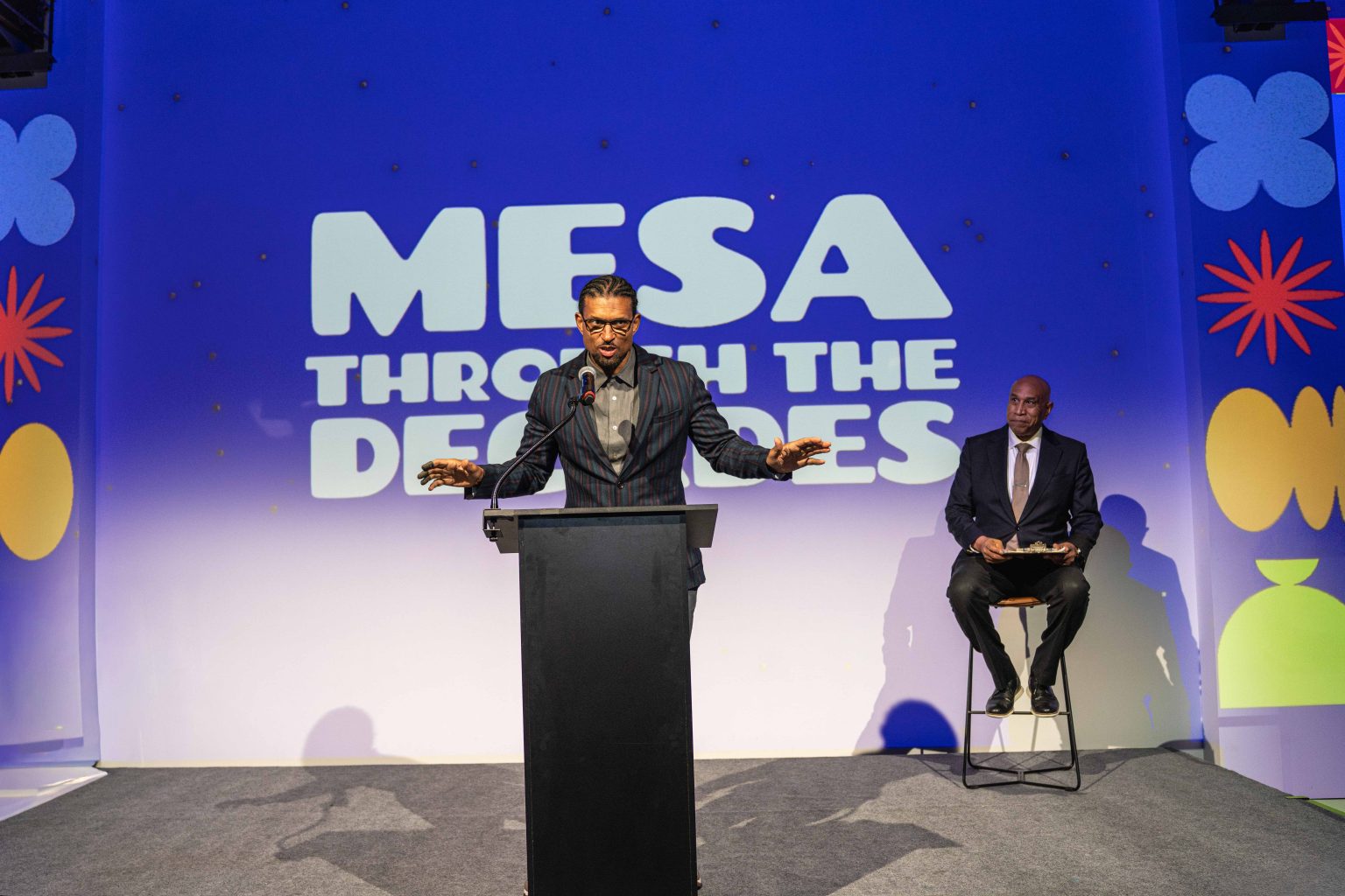 A man speaks at a podium on stage with "MESA Through the Decades 2025 Photo Gallery" projected behind him. Another man in a suit sits on a tall stool to the right, against a background of colorful, abstract shapes.