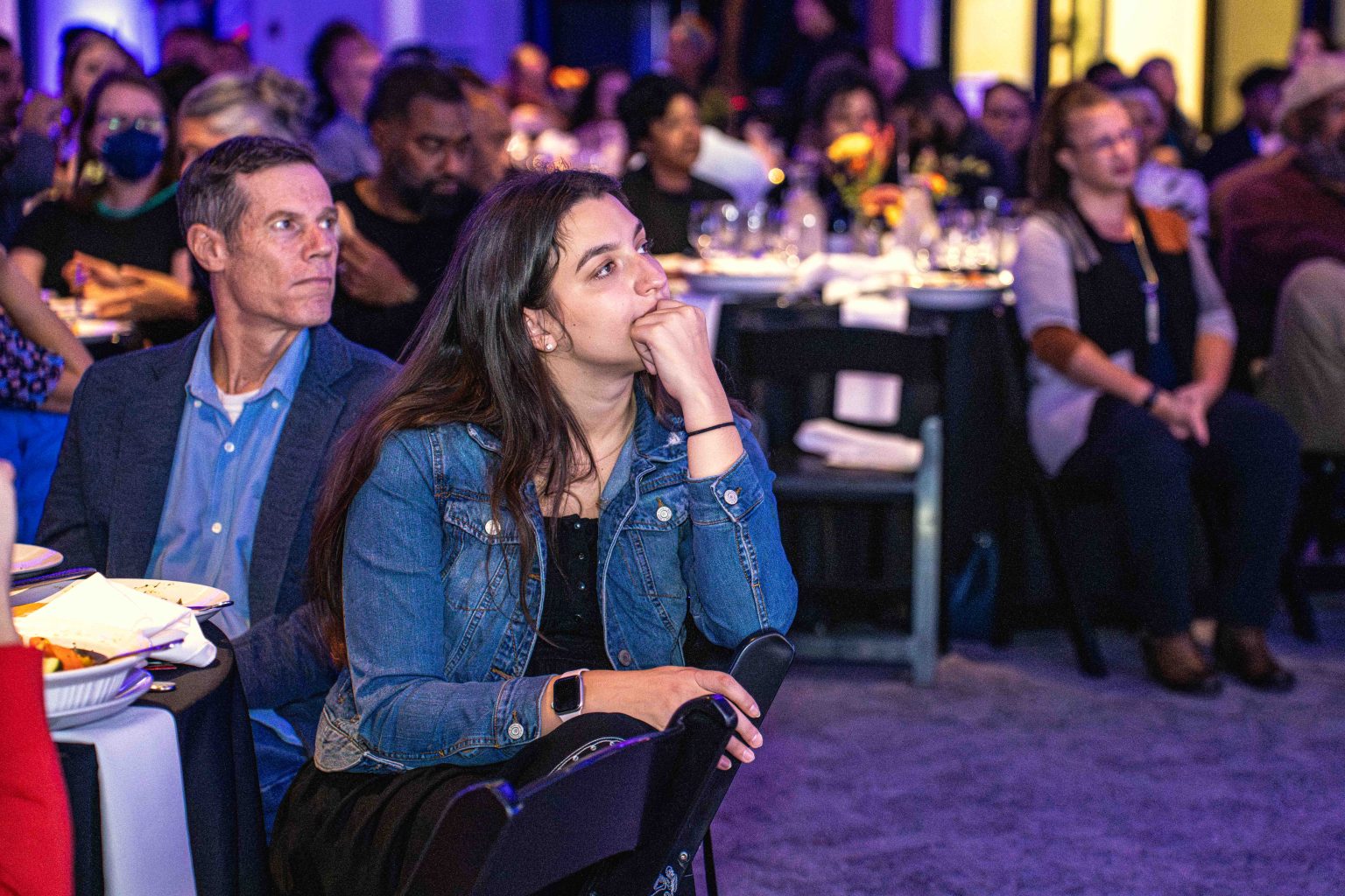 A group of people sit at round tables, attentively watching something off-camera. In this MESA Through the Decades 2025 Photo Gallery, a woman in the foreground rests her chin on her hand as others listen quietly in a dimly lit event space.