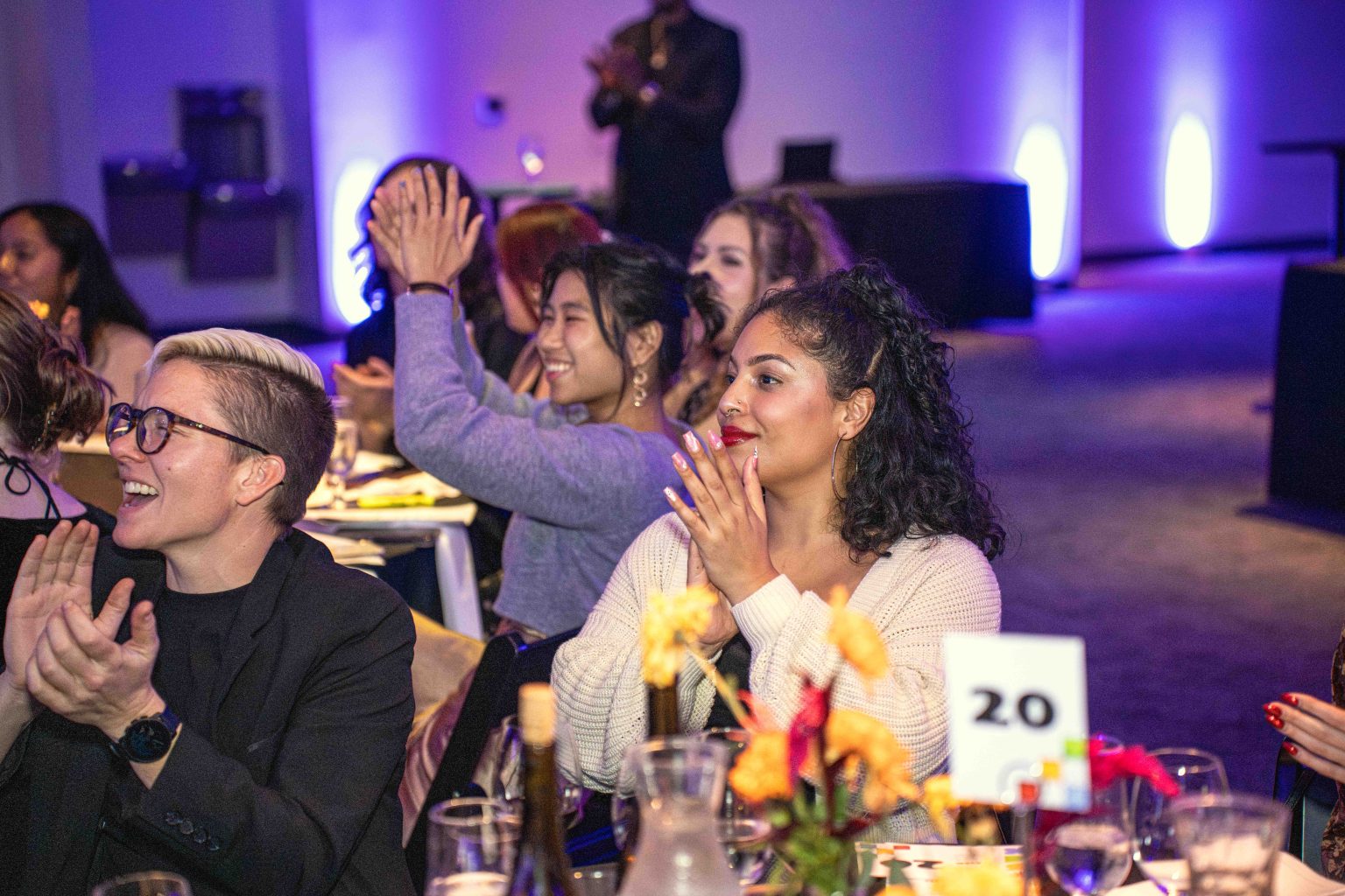 A group of people sitting at a decorated table clap and smile during the MESA Through the Decades 2025 Photo Gallery event in a dimly lit room with purple lighting and flower centerpieces.