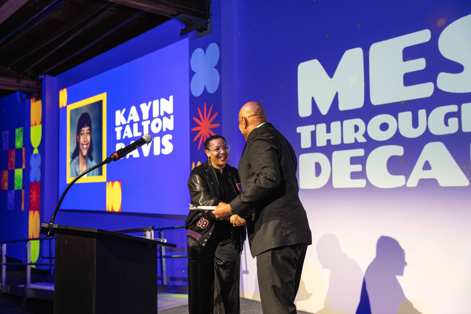 Two people stand on stage shaking hands and smiling under bright lights. A large screen behind them displays MESS THROUGH DECA and Kayin Talton Davis, with a photo and colorful graphics from the MESA Through the Decades 2025 Photo Gallery.
