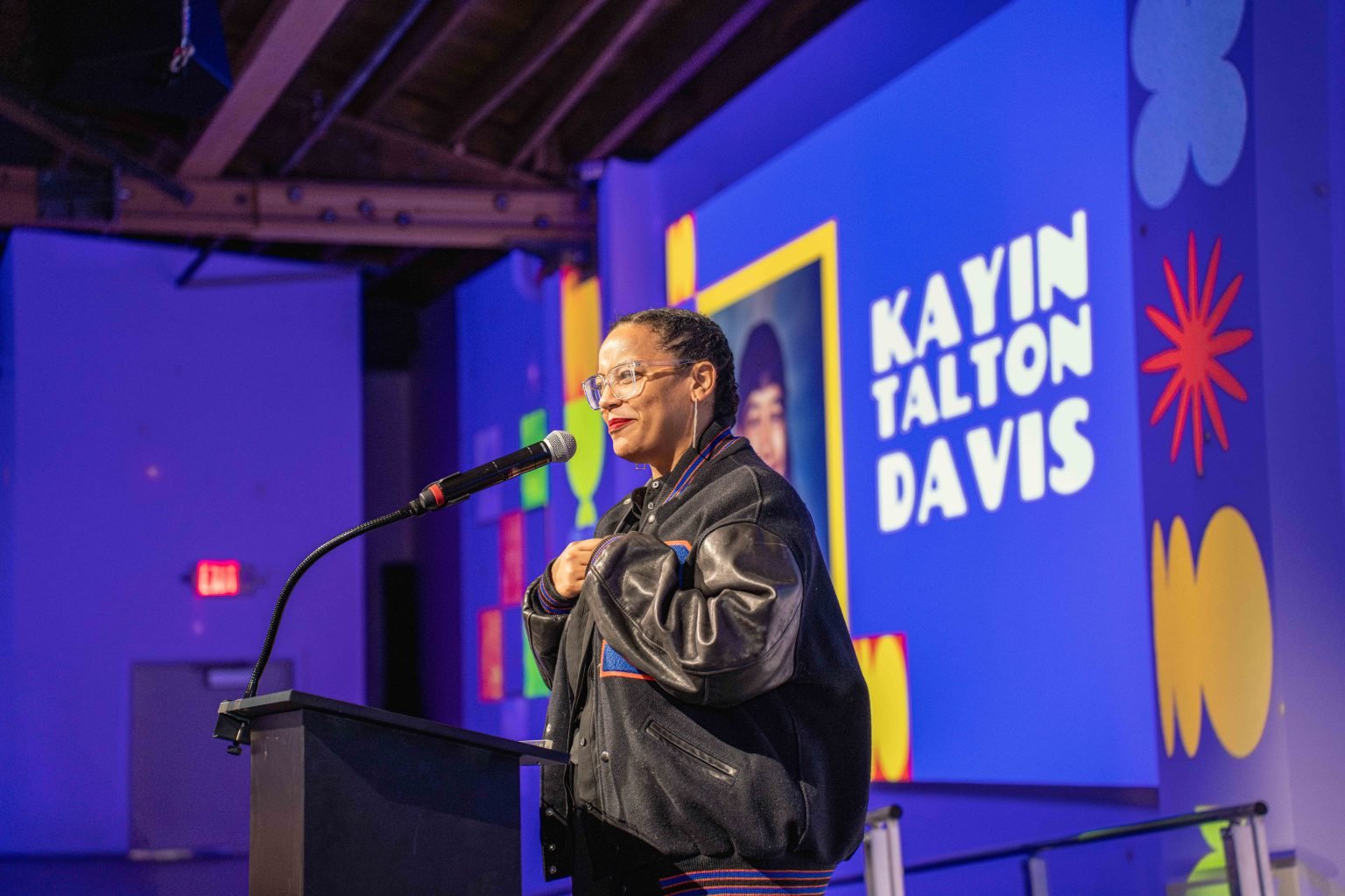 A person wearing glasses and a leather jacket speaks at a podium, with a colorful screen behind them displaying the name Kayin Talton Davis and various graphic shapes, captured for the MESA Through the Decades 2025 Photo Gallery.