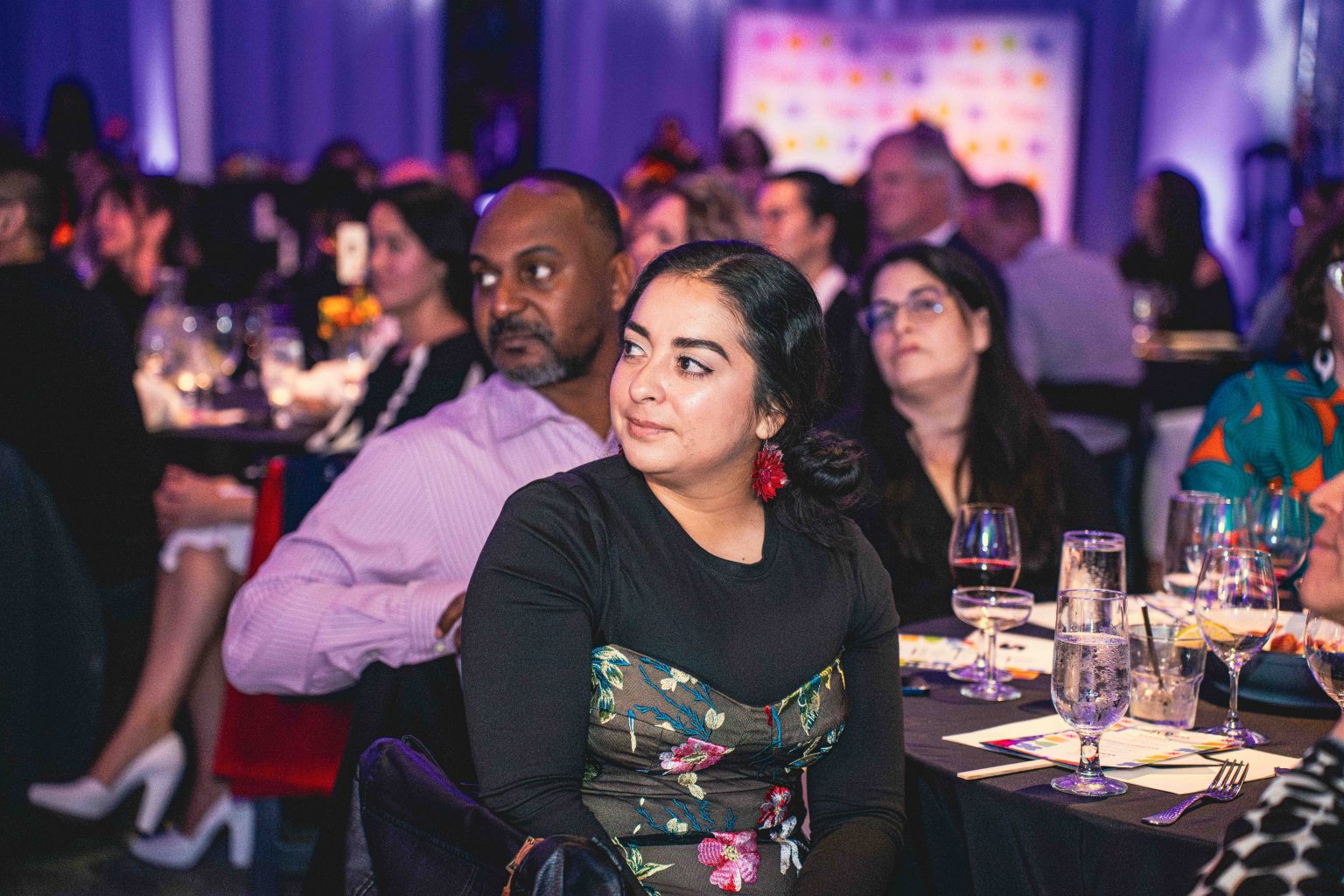 A woman in a black floral dress sits at a table, attentively watching an event at the MESA Through the Decades 2025 Photo Gallery. She is surrounded by other guests in formal attire, with dim lighting and a purple-lit background.