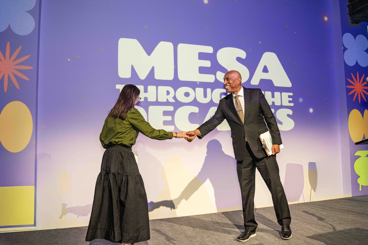 A woman and a man in formal attire shake hands on stage in front of a screen displaying MESA THROUGH THE DECADES. Both are smiling as the man holds a folder, captured for the MESA Through the Decades 2025 Photo Gallery.