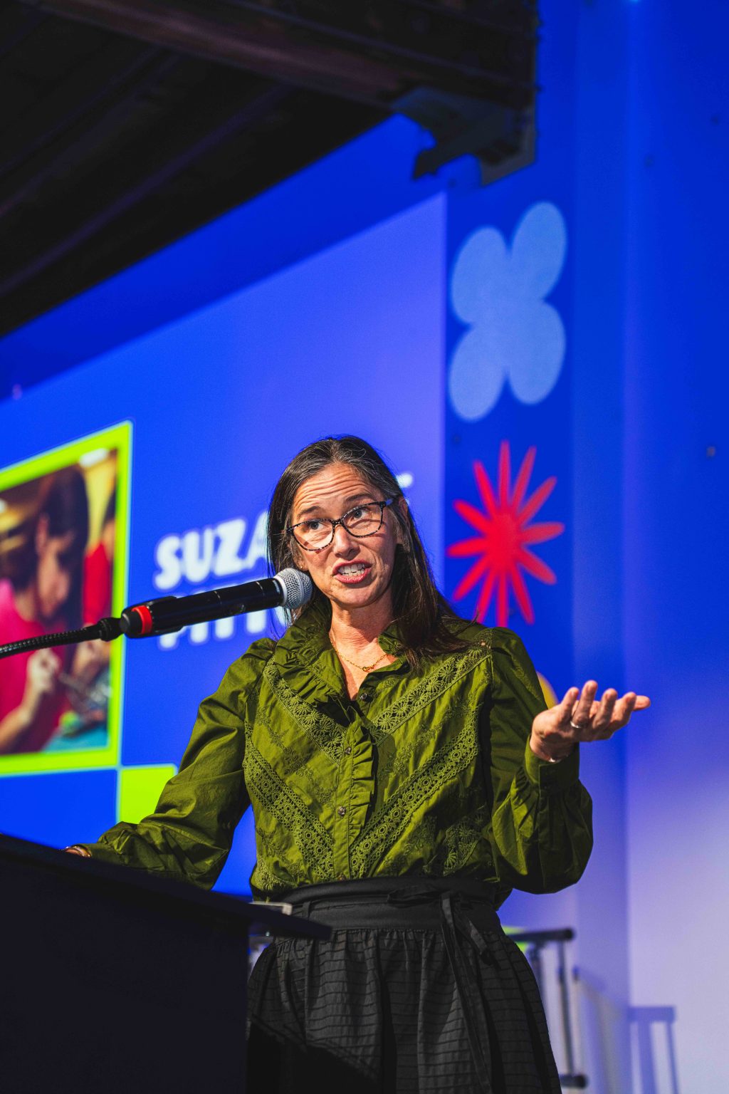 A woman with long brown hair and glasses speaks at a podium, gesturing with one hand. She wears a green blouse and stands before the colorful blue backdrop of the MESA Through the Decades 2025 Photo Gallery.