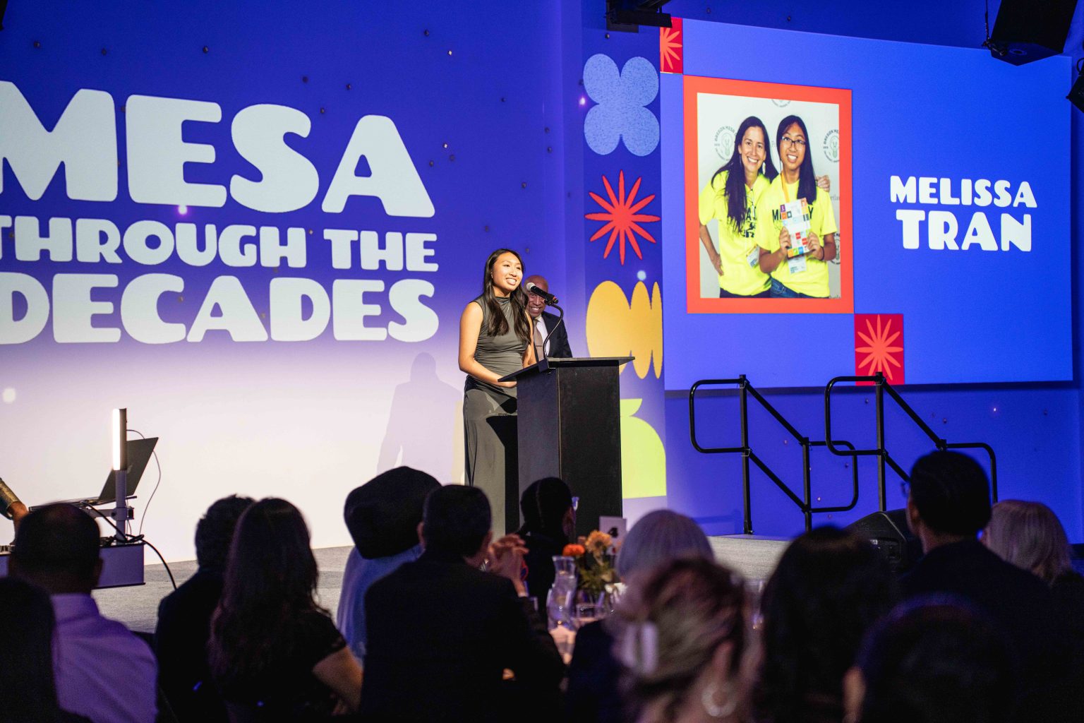 A woman speaks at a podium on stage during the MESA Through the Decades 2025 Photo Gallery event, with images and Melissa Tran’s name projected on a large screen as audience members watch.