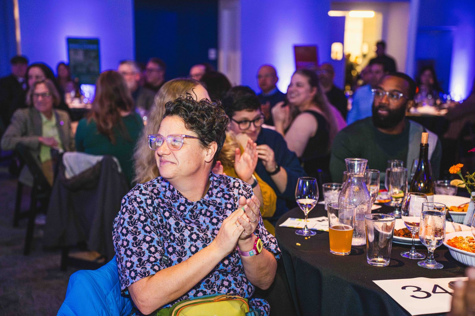 A group of people sit at round tables in a dimly lit event space, clapping and smiling. Glasses, bottles, and plates are on the tables as blue and purple lighting creates a festive atmosphere at the MESA Through the Decades 2025 Photo Gallery.