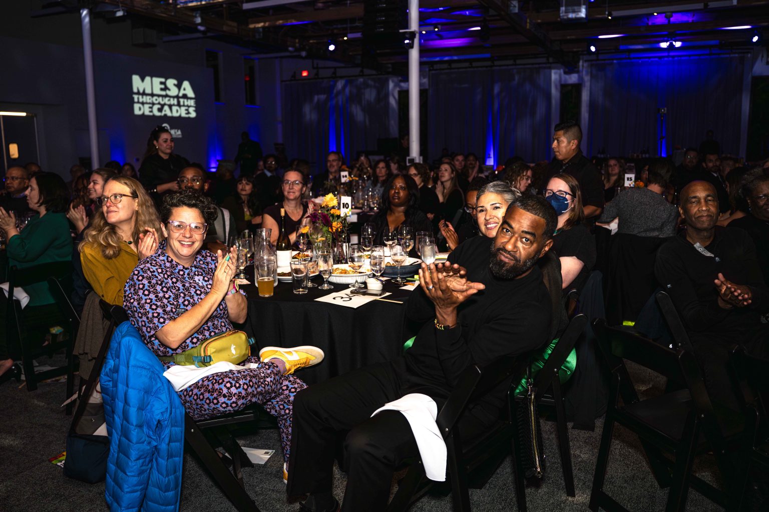 A diverse group of people sit around tables at a formal event, clapping and smiling. Projected text on the wall reads, “MESA THROUGH THE DECADES.” The MESA Through the Decades 2025 Photo Gallery captures this vibrant, blue-lit celebration.