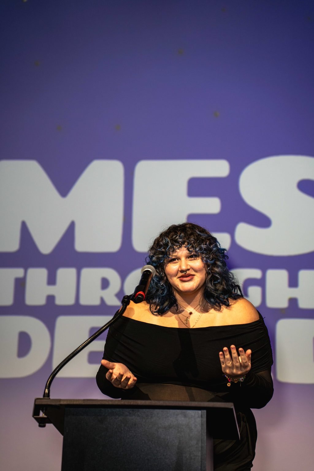A person with curly blue hair and an off-the-shoulder black top speaks at a podium with a microphone, in front of a blurred purple background featuring large white text from the MESA Through the Decades 2025 Photo Gallery.
