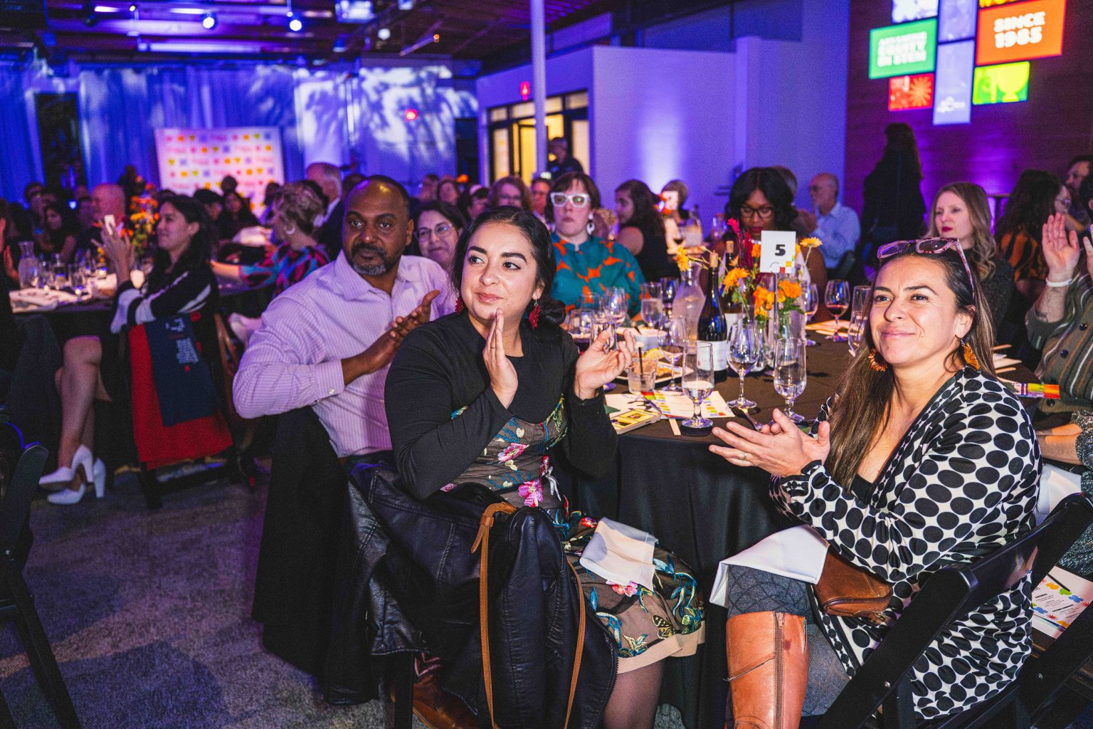 A diverse group of people sit at round tables, clapping and smiling at an indoor event, with colorful lights and artwork on the walls in the background—a moment captured for the MESA Through the Decades 2025 Photo Gallery.