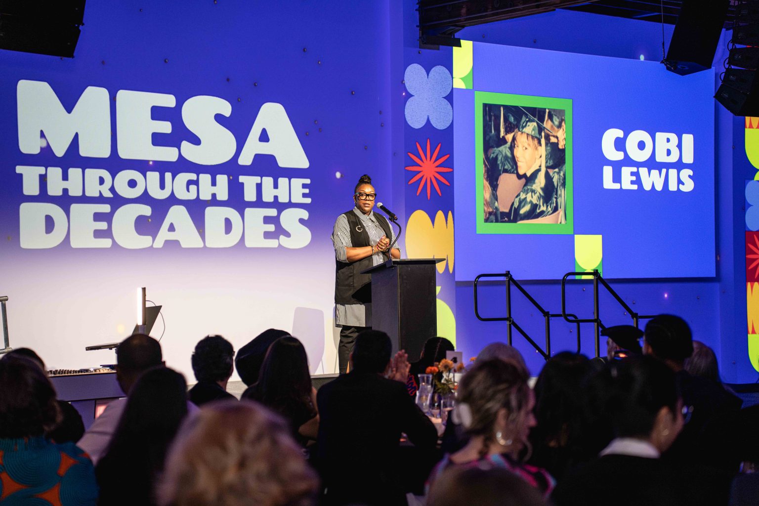 A speaker stands at a podium onstage at an event with a screen displaying MESA THROUGH THE DECADES and a picture labeled COBI LEWIS, as the audience watches attentively—captured in the MESA Through the Decades 2025 Photo Gallery.