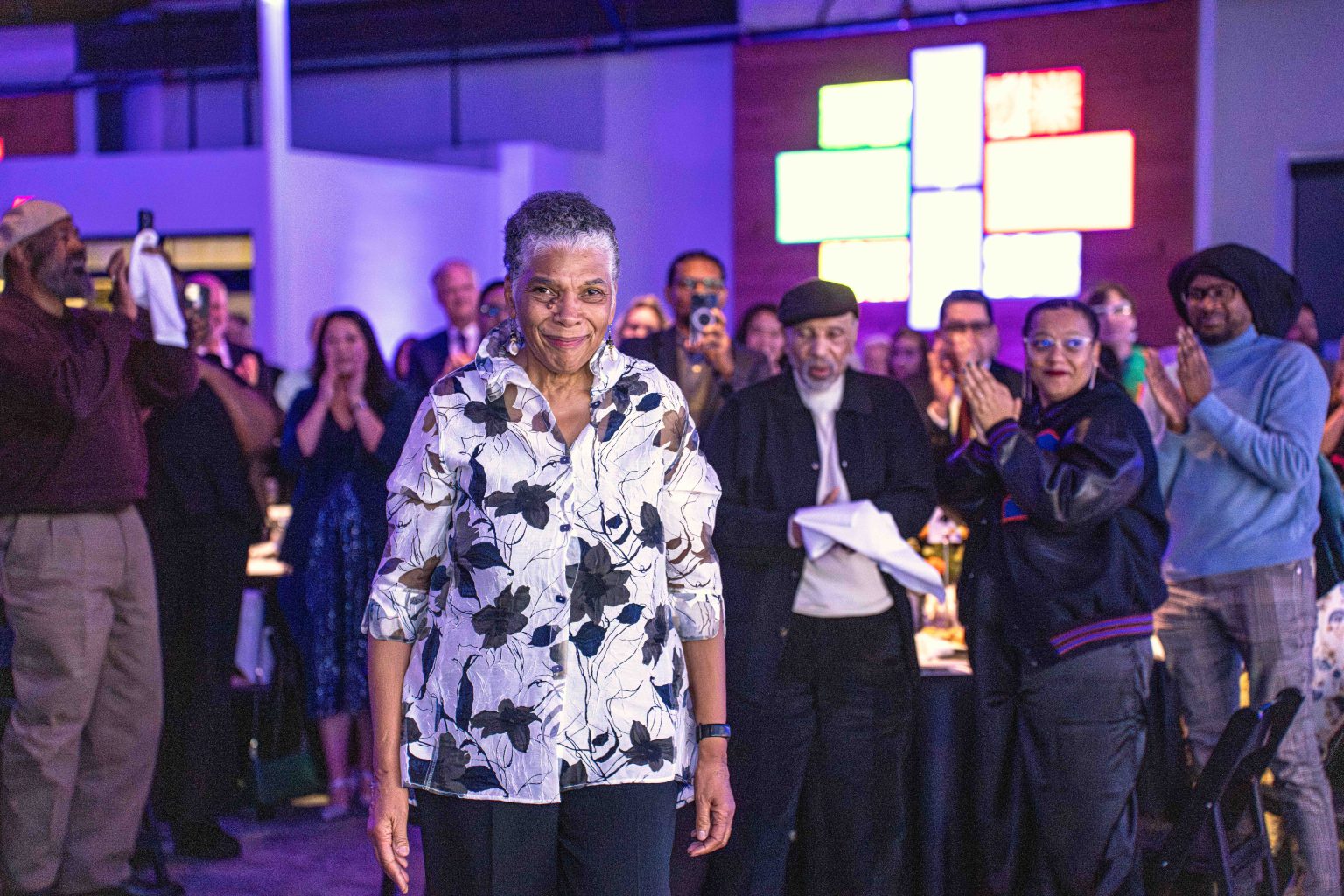 An older woman in a white and black floral shirt stands smiling as people clap and cheer for her in an indoor event space with colorful lights, captured at the MESA Through the Decades 2025 Photo Gallery.