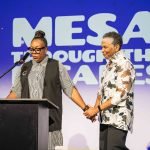 Two women stand on stage holding hands; one speaks at a podium while the other smiles. Behind them, large text reads, MESA THROUGH THE AGES. The setting appears to be an event, part of the MESA Through the Decades 2025 Photo Gallery.
