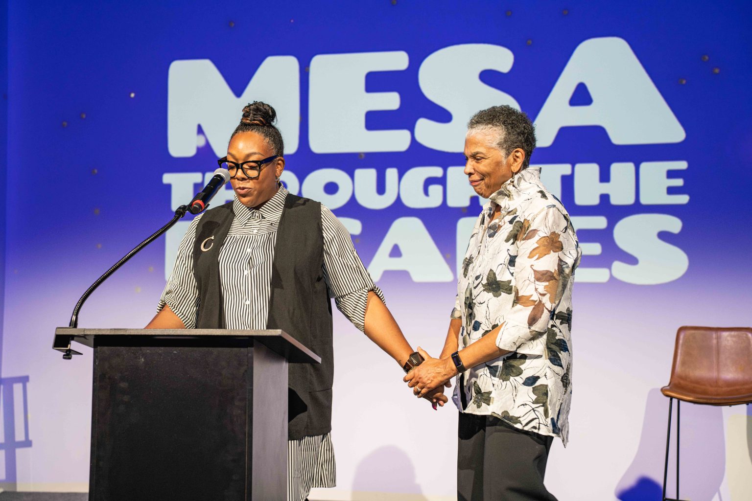Two women stand on stage holding hands; one speaks at a podium while the other smiles. Behind them, large text reads, MESA THROUGH THE AGES. The setting appears to be an event, part of the MESA Through the Decades 2025 Photo Gallery.