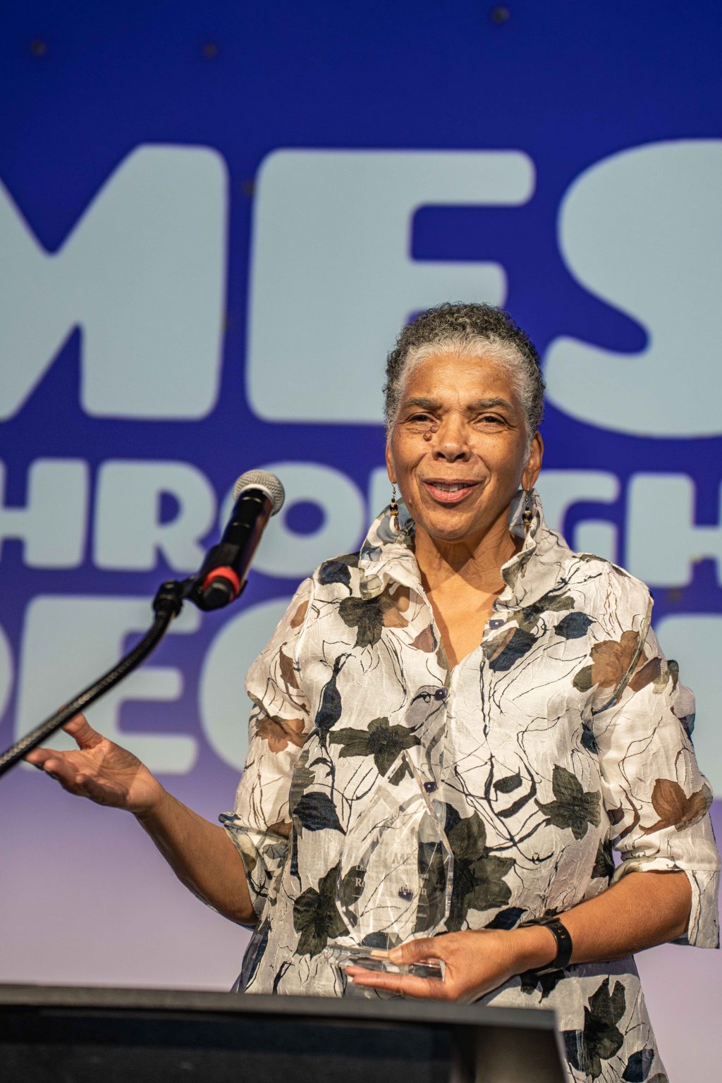 An older woman with short gray hair and hoop earrings speaks at a microphone. Smiling in a floral blouse, she stands before a blue backdrop with large white letters for the MESA Through the Decades 2025 Photo Gallery.