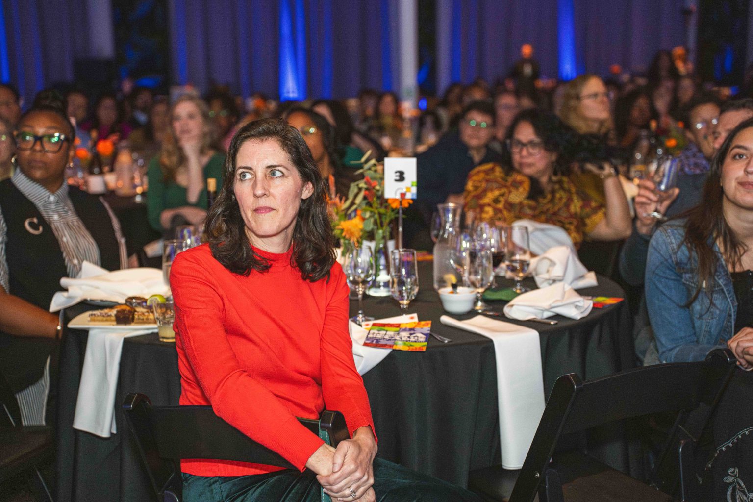 A woman in a red top sits at a table, looking to her left, amid the crowd at the formal MESA Through the Decades 2025 Photo Gallery event with decorated tables, glasses, and floral centerpieces. Other attendees are seated and watching attentively.
