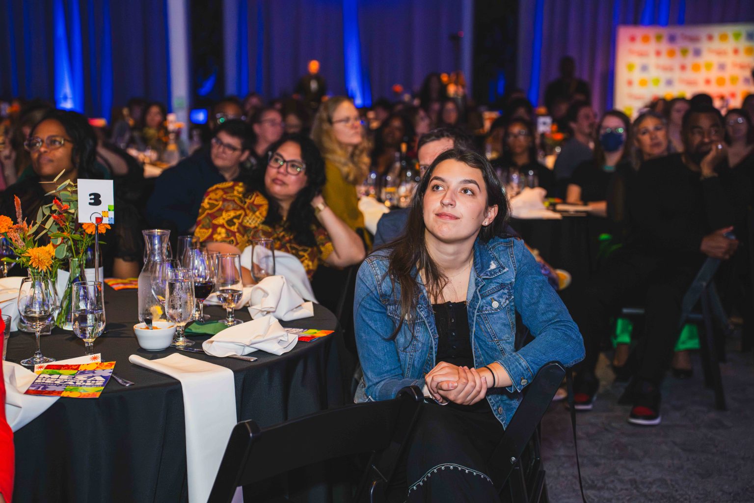 At the MESA Through the Decades 2025 Photo Gallery, a young woman in a denim jacket sits at a round table, attentively watching an event on stage amid guests at decorated tables in a colorful, warmly lit venue.