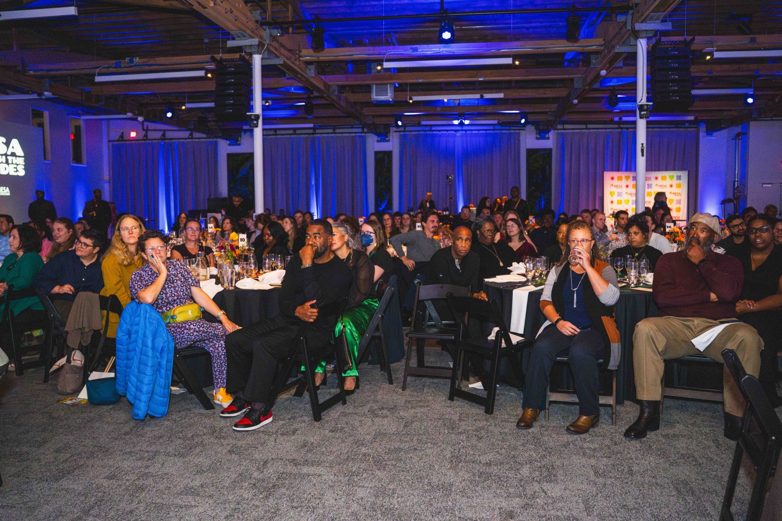 A diverse audience sits at round tables, attentively watching an event in a modern, dimly lit venue with blue and purple lighting. This MESA Through the Decades 2025 Photo Gallery captures people of all ages engaged and interested in the program.