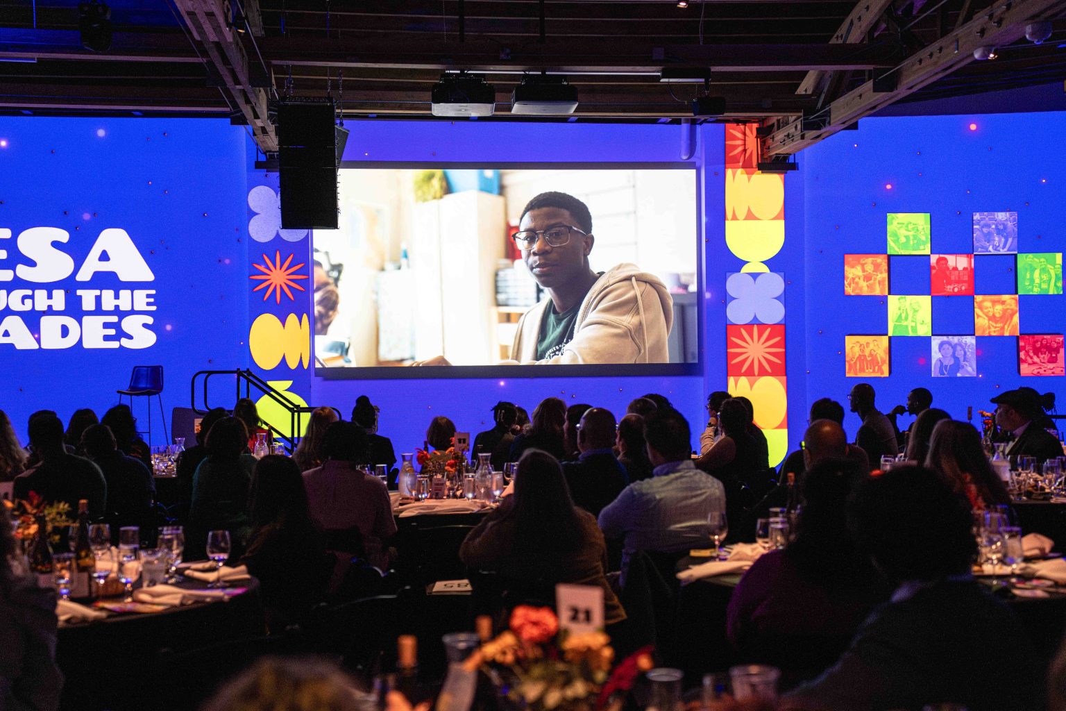 A crowd sits at tables in a dimly lit event space, watching a large screen displaying a man in glasses. Colorful graphics and the words MESA Through the Decades 2025 Photo Gallery appear on the blue-lit walls.