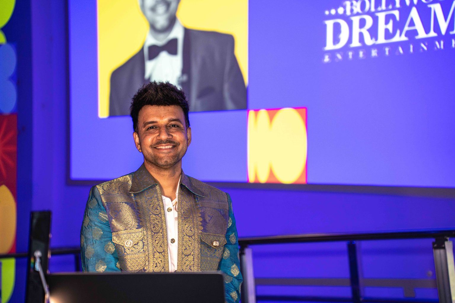 A man with dark hair, wearing a blue and gold patterned jacket, smiles while standing in front of a laptop. Behind him, the screen displays a DREAM message and highlights from the MESA Through the Decades 2025 Photo Gallery.