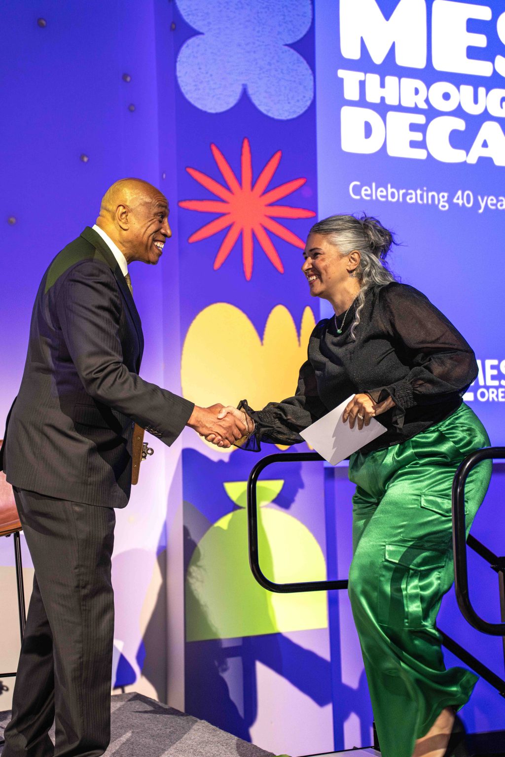 A man in a dark suit and a woman in a black blouse and shiny green pants shake hands and smile on stage at an event, with colorful floral graphics and "Celebrating 40 years" and MESA Through the Decades 2025 Photo Gallery in the background.